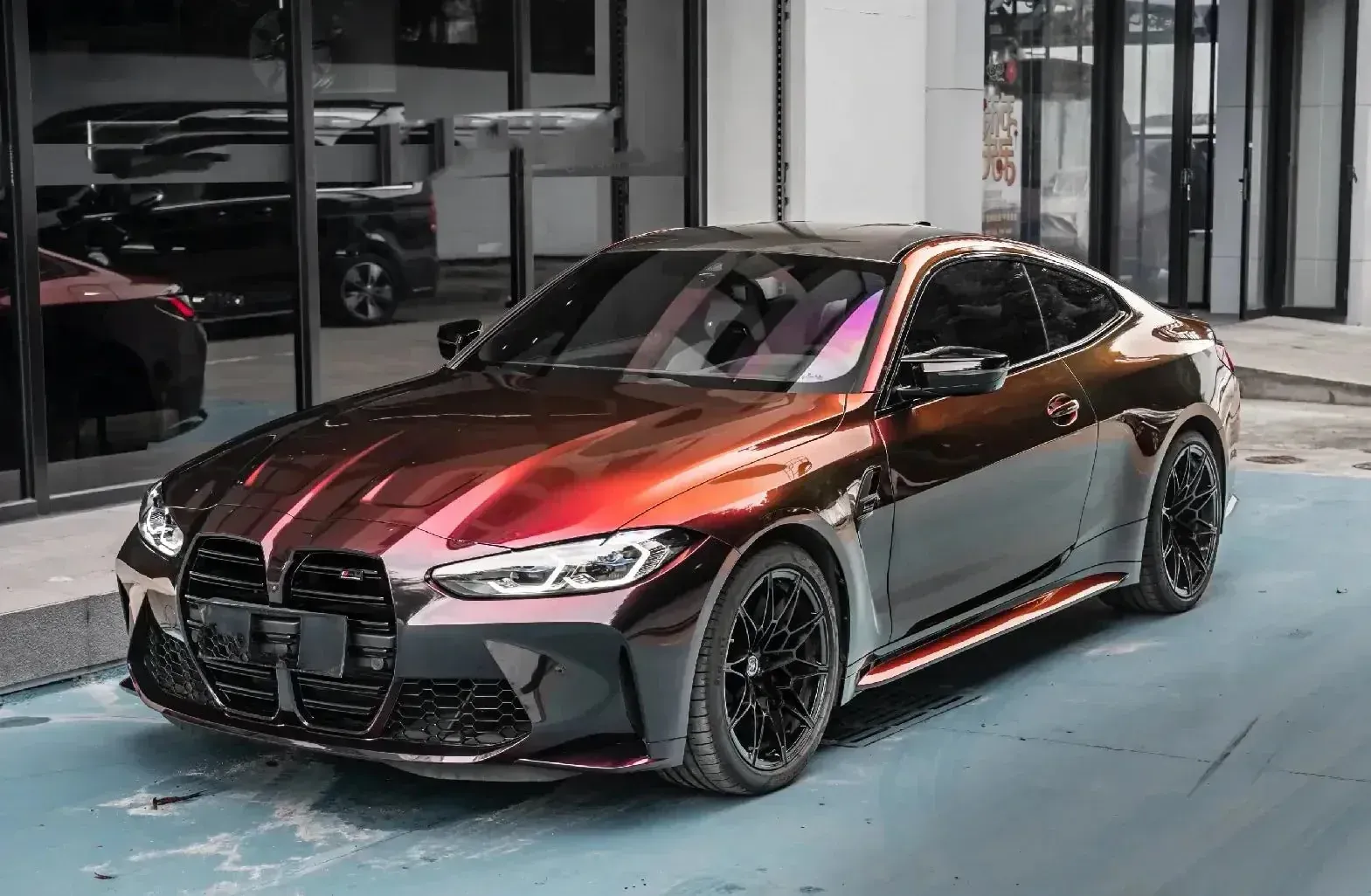 A shiny, color-shifting BMW M4 coupe parked outside. Dark red to black, black wheels, modern storefront.