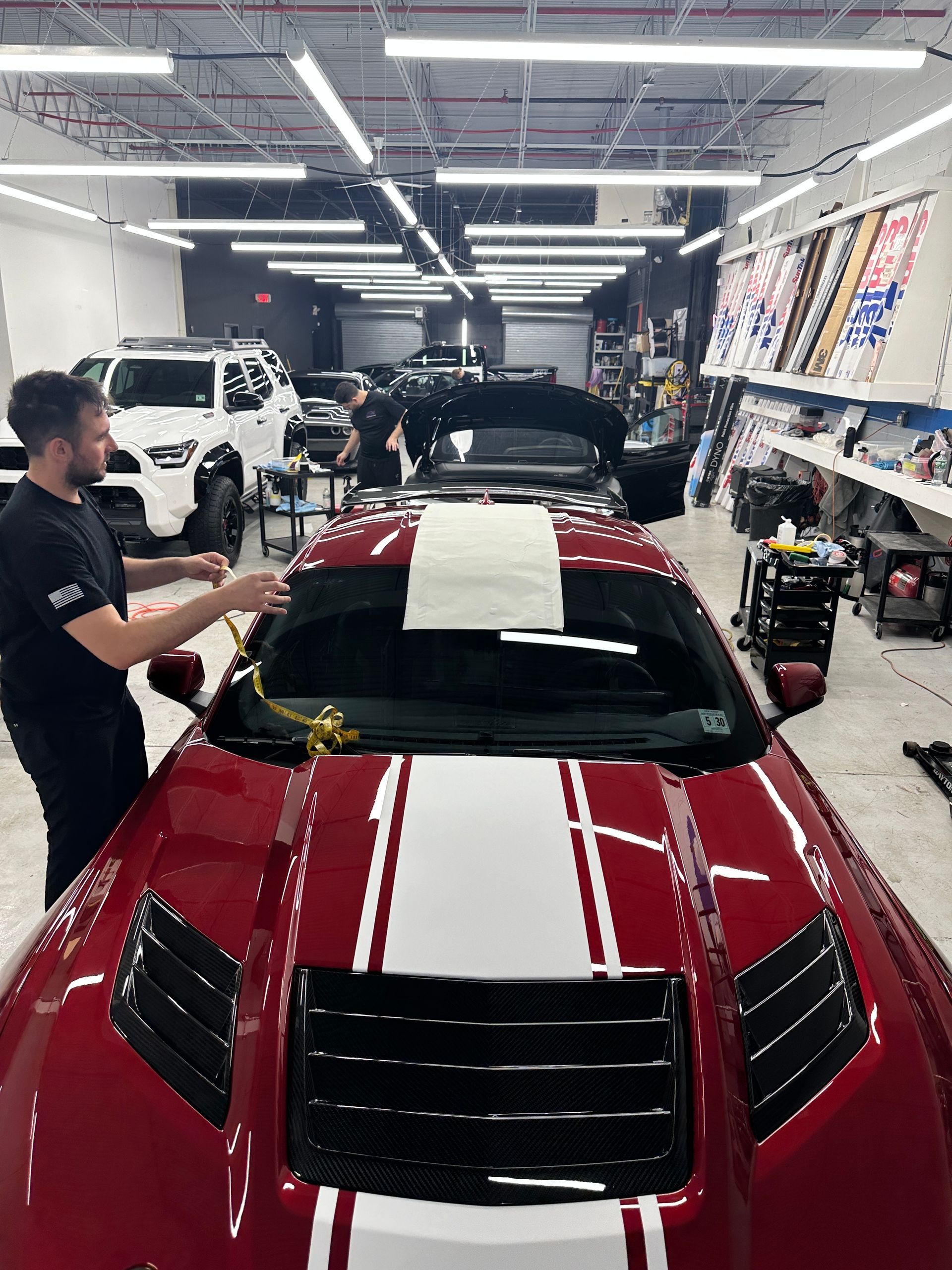 A worker applying film to a red sports car in a shop. Bright overhead lights, other cars and equipment visible.