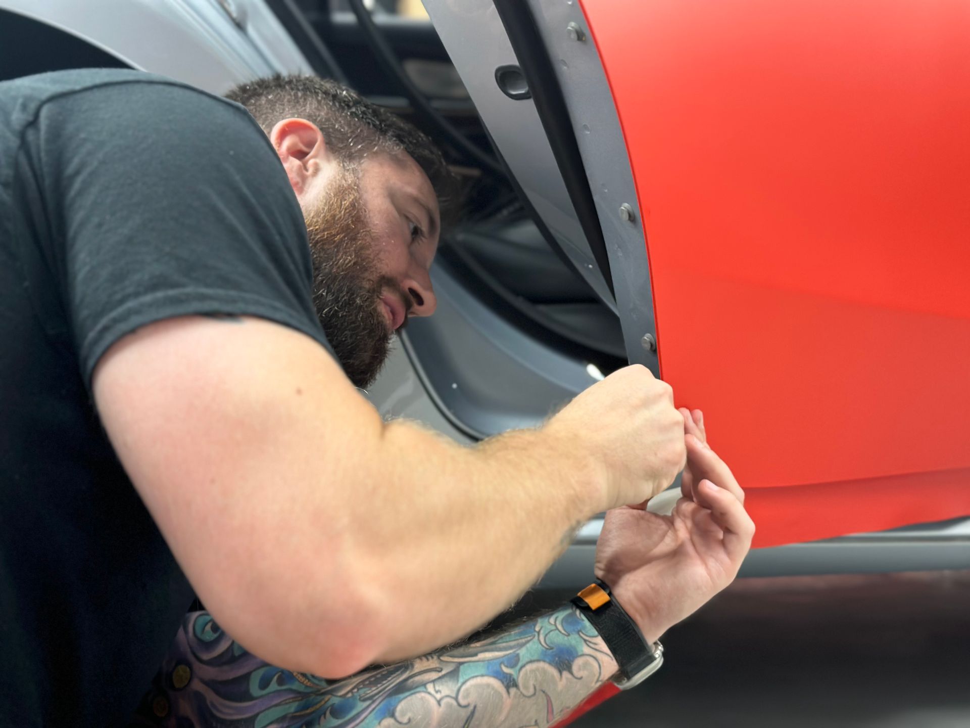 Man installing parts on a car door, wearing a black shirt with tattoos on his arm.