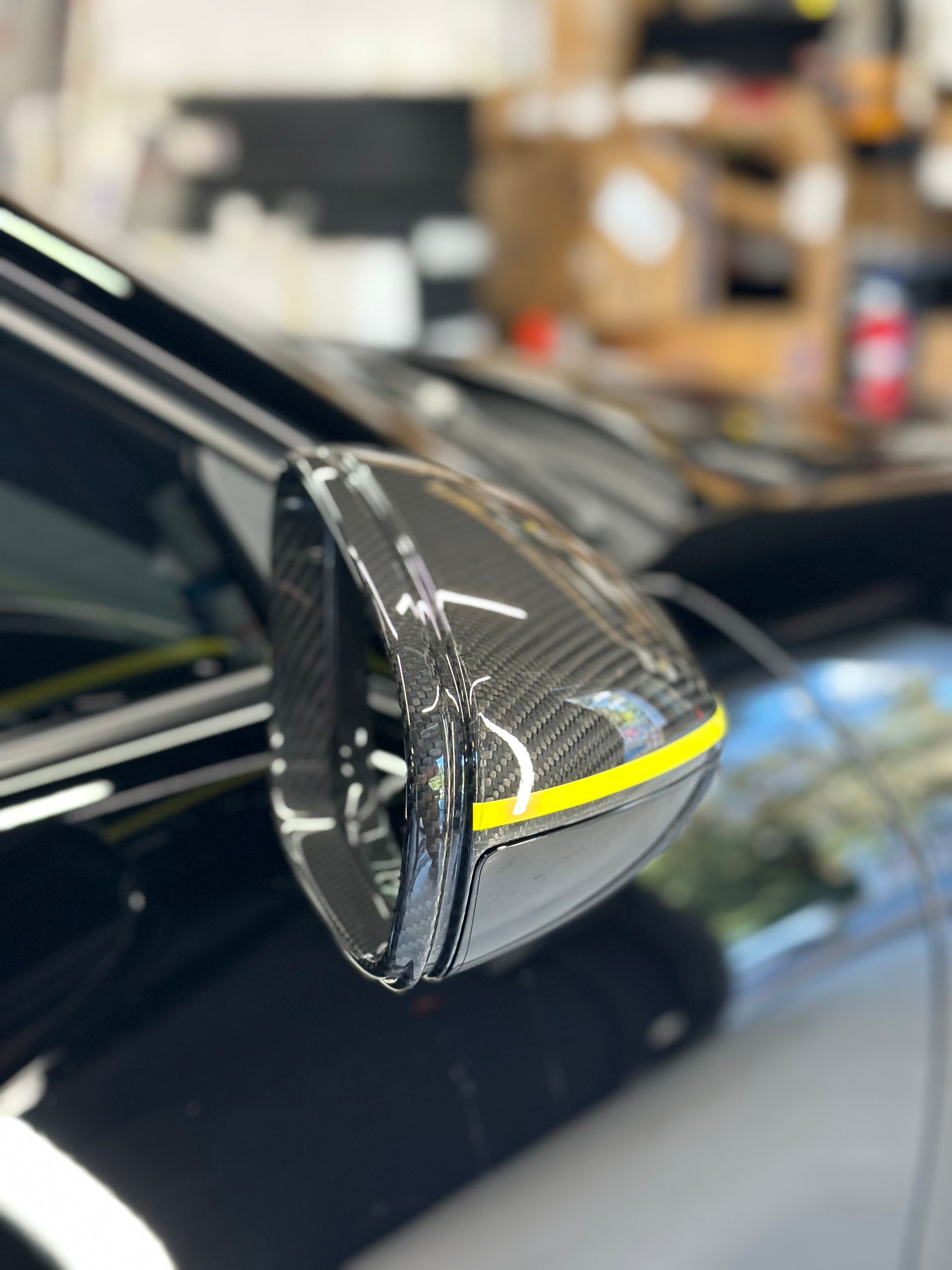 Carbon fiber side mirror with yellow stripe on a black car in a shop.