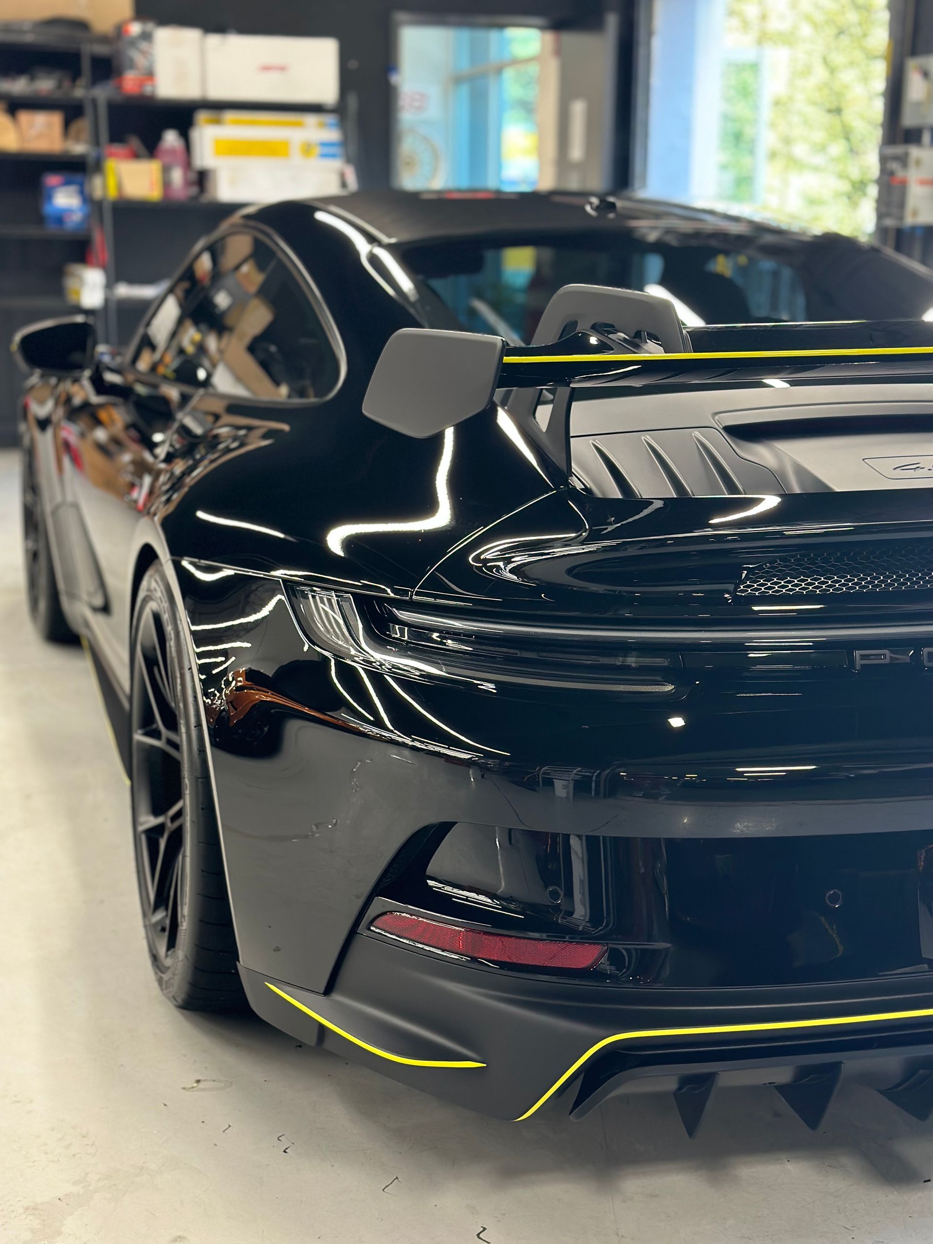 Close-up of a black car's side mirror with carbon fiber details and a yellow stripe, set in a workshop environment.