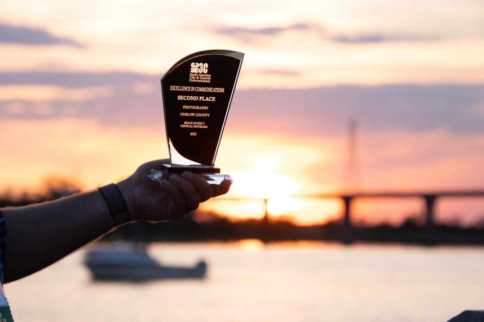 A hand holding an NC3C Excellence in Communications Award trophy at sunset during the 2023 Wilmington conference