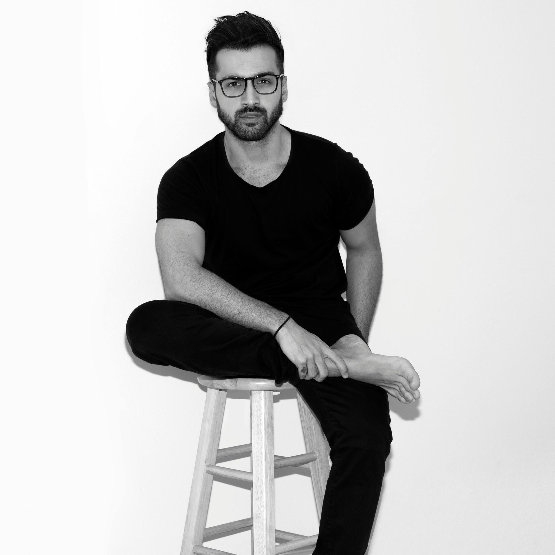 A man with glasses is sitting on a stool with his legs crossed