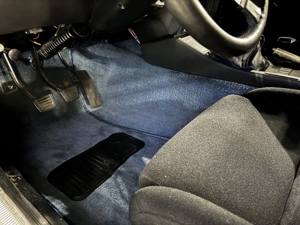 Interior car shot: blue carpet, black seat, pedals, and steering wheel.