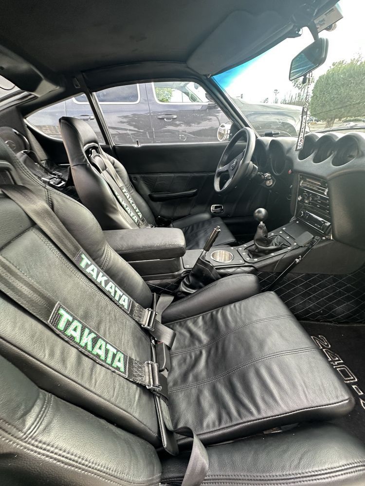 Black interior of a vintage sports car, with Takata racing harnesses, seats, and a manual transmission.