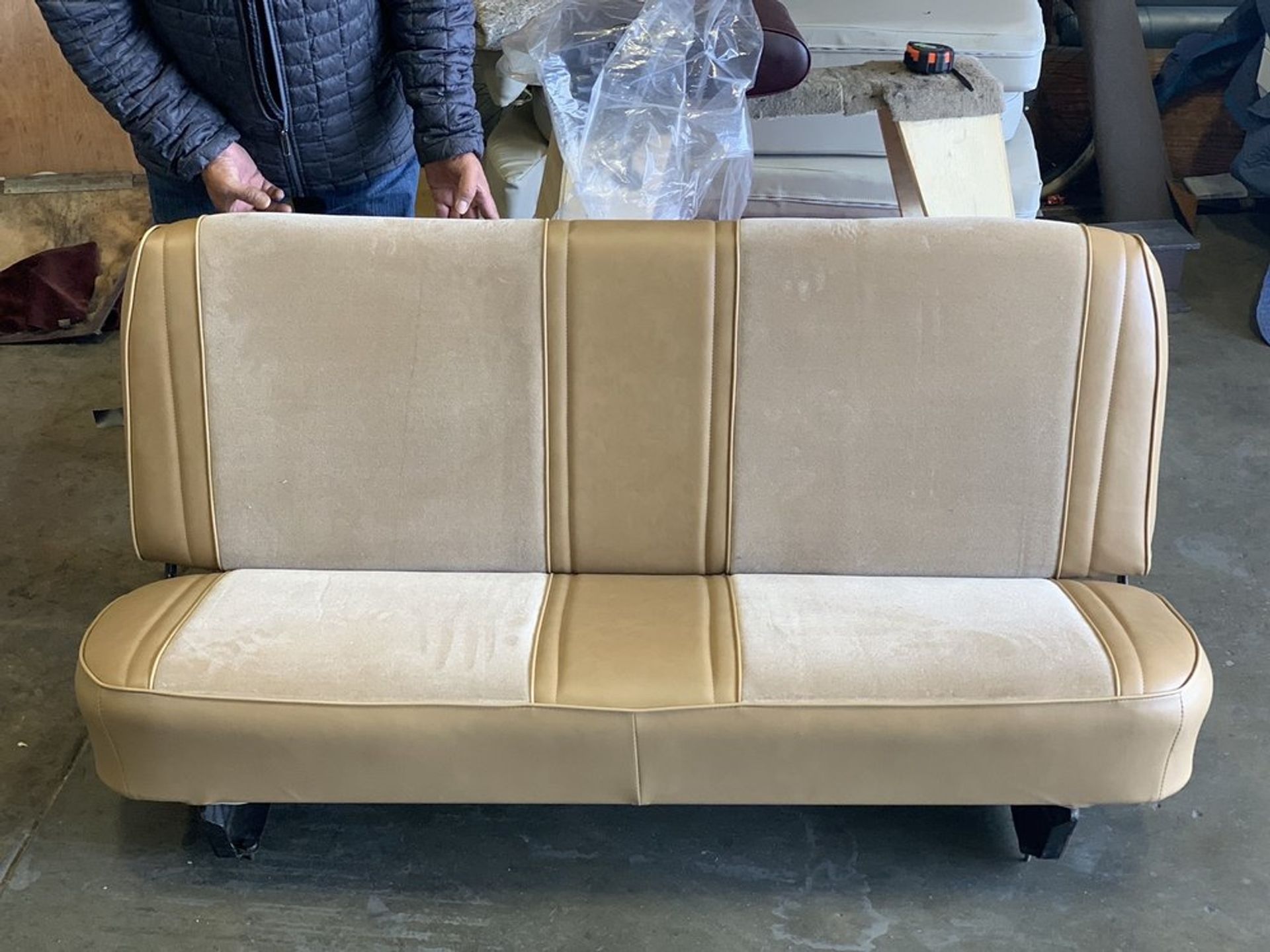 Beige and tan vehicle bench seat held by a person in a workshop.