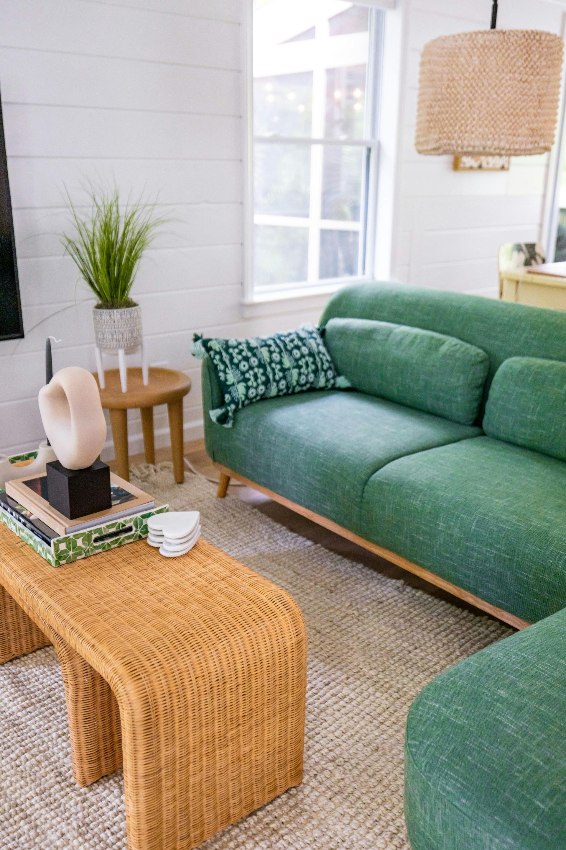 Green sofa in a cozy living room, with wicker coffee table, plants, and woven light fixture.