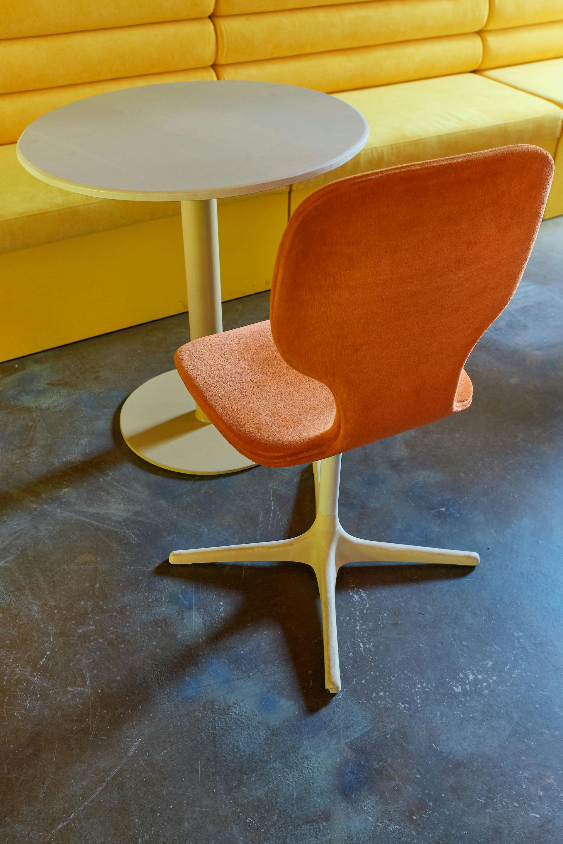 Orange chair in front of a small round table, yellow seating in the background, on a dark gray floor.