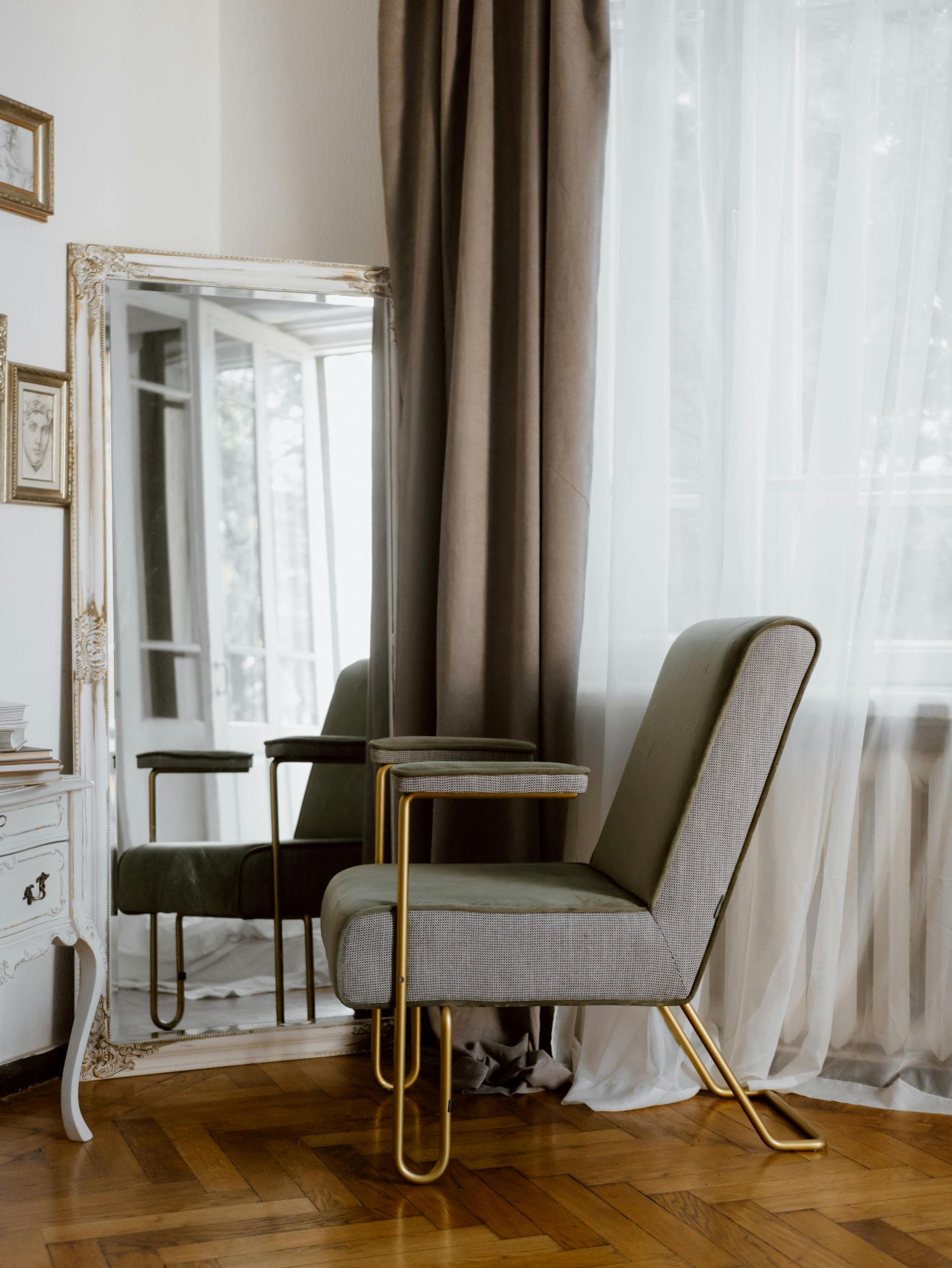 A stylish armchair with gold legs and small side tables stands in front of a large mirror and window with curtains.