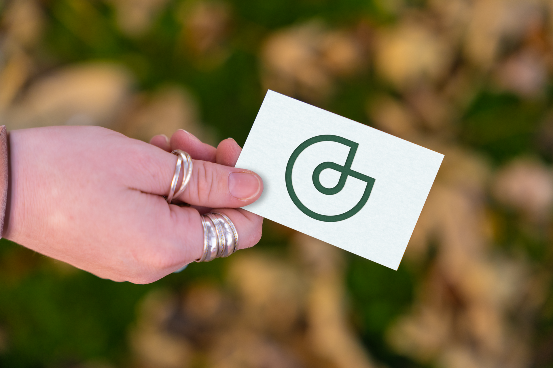 Main tenant une carte de visite blanche avec un logo vert, sur un fond naturel flou.