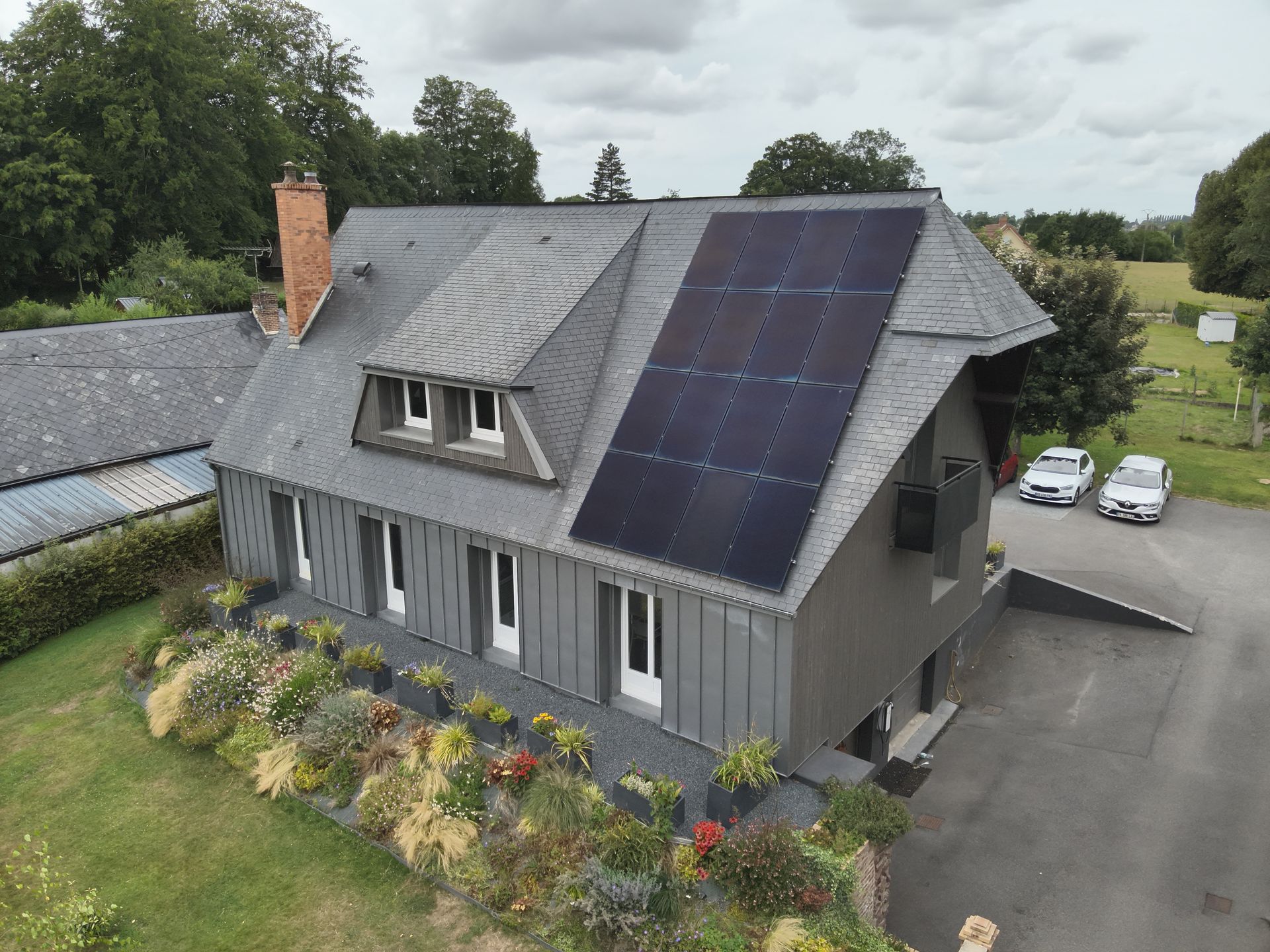 Une maison gris foncé avec un panneau solaire sur le toit, des fleurs devant et des voitures garées à proximité.