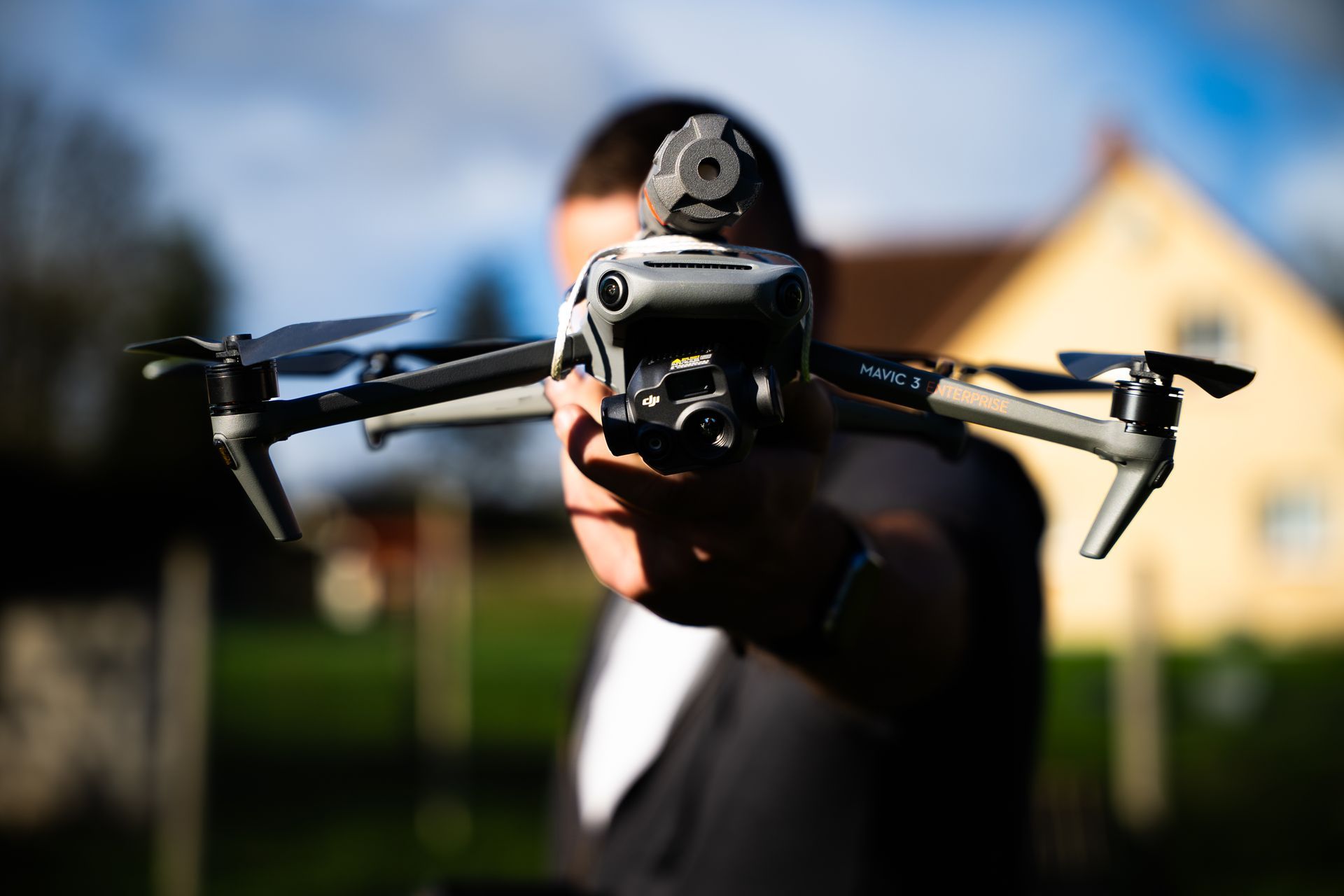 Une personne tient un drone face à la caméra. La caméra du drone est visible. Paysage extérieur ensoleillé avec un bâtiment 