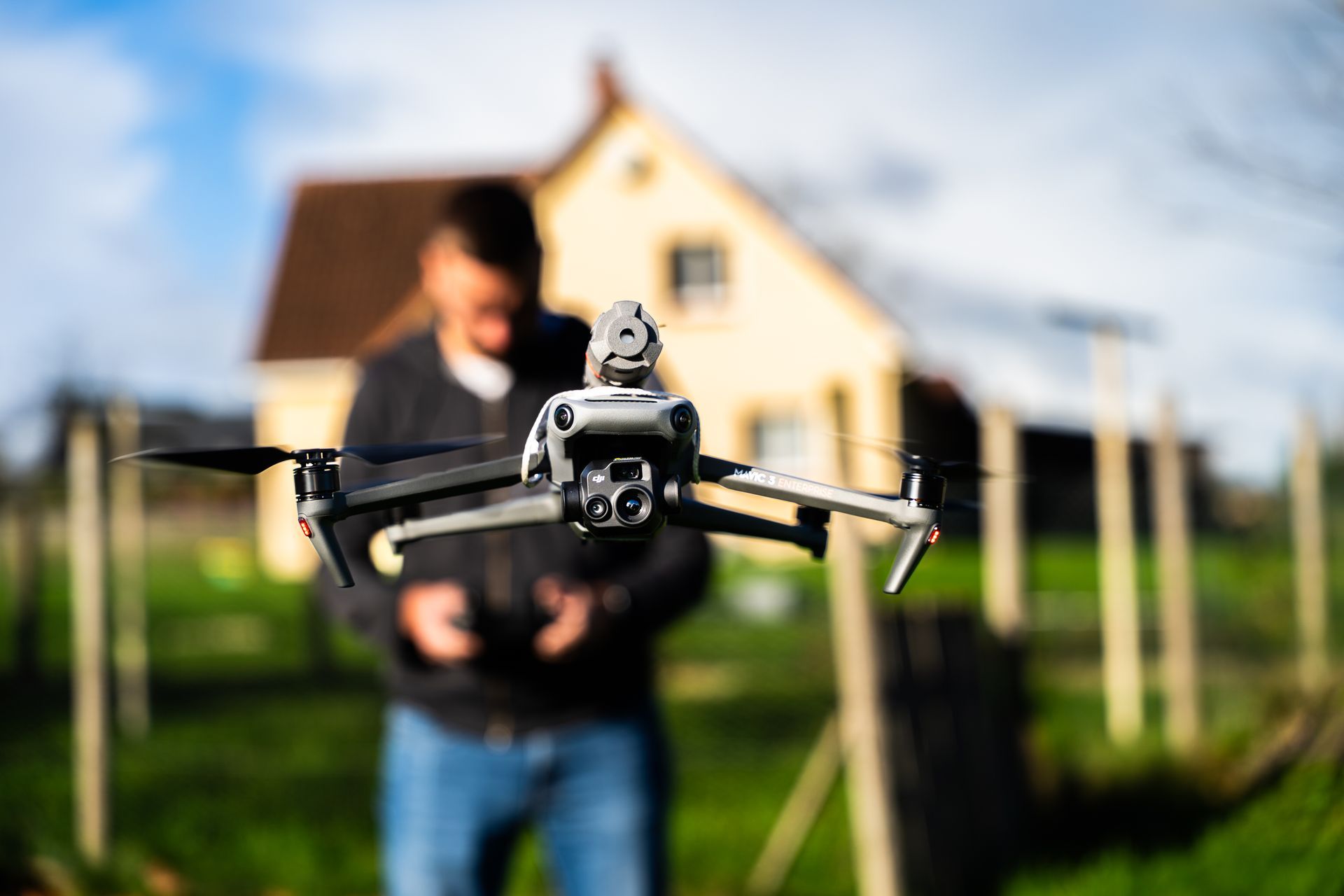 Drone en vol, piloté par une personne floue, devant une maison.