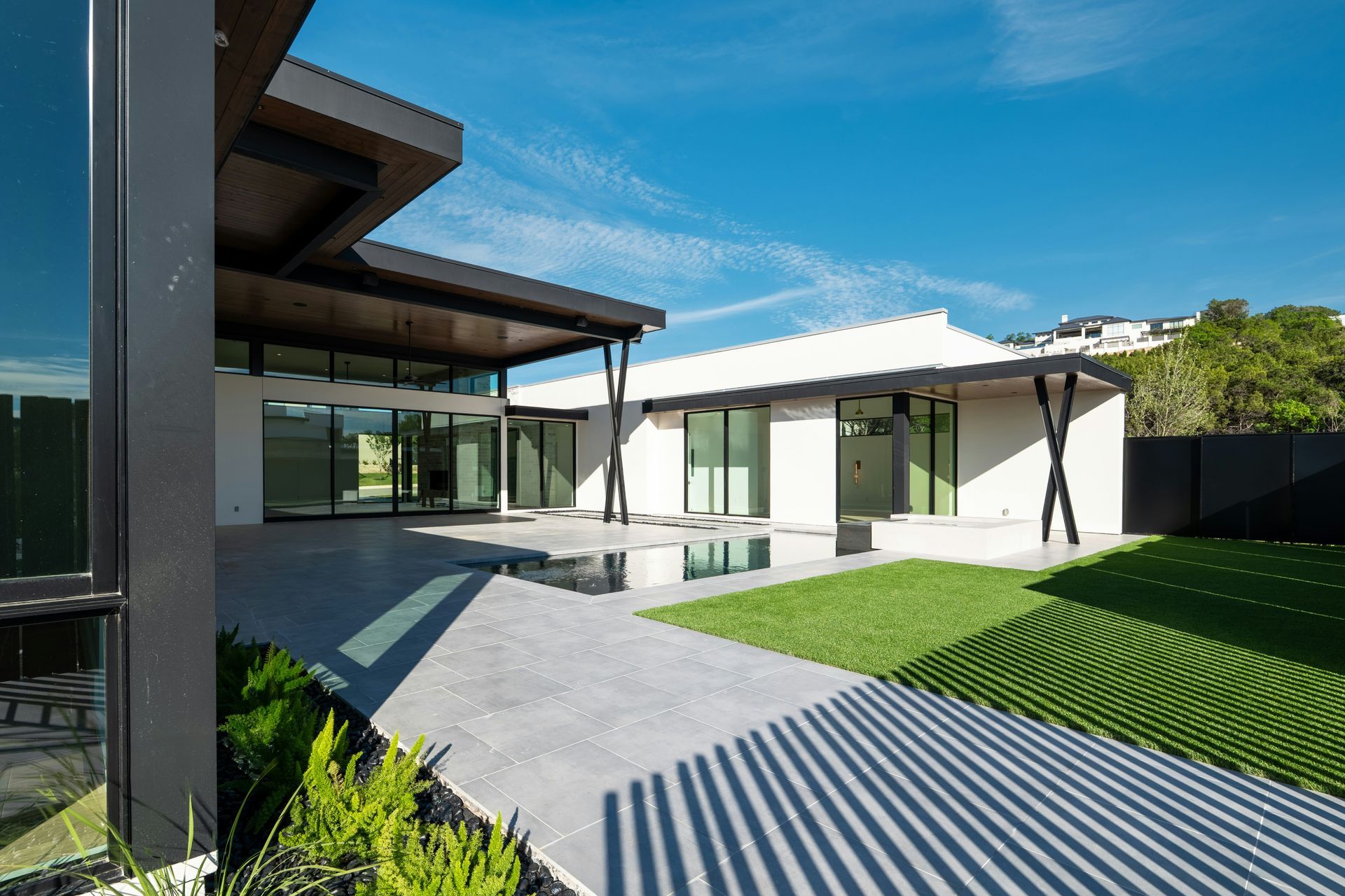 Modern house courtyard with white walls, glass doors, gray paving, and a green lawn under a blue sky