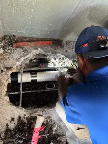 A professional in a blue shirt and baseball cap works on a white PVC pipe in an outdoor trench.