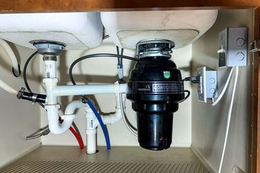 A kitchen sink cabinet interior featuring white PVC plumbing pipes, a black garbage disposal, and electrical wiring.