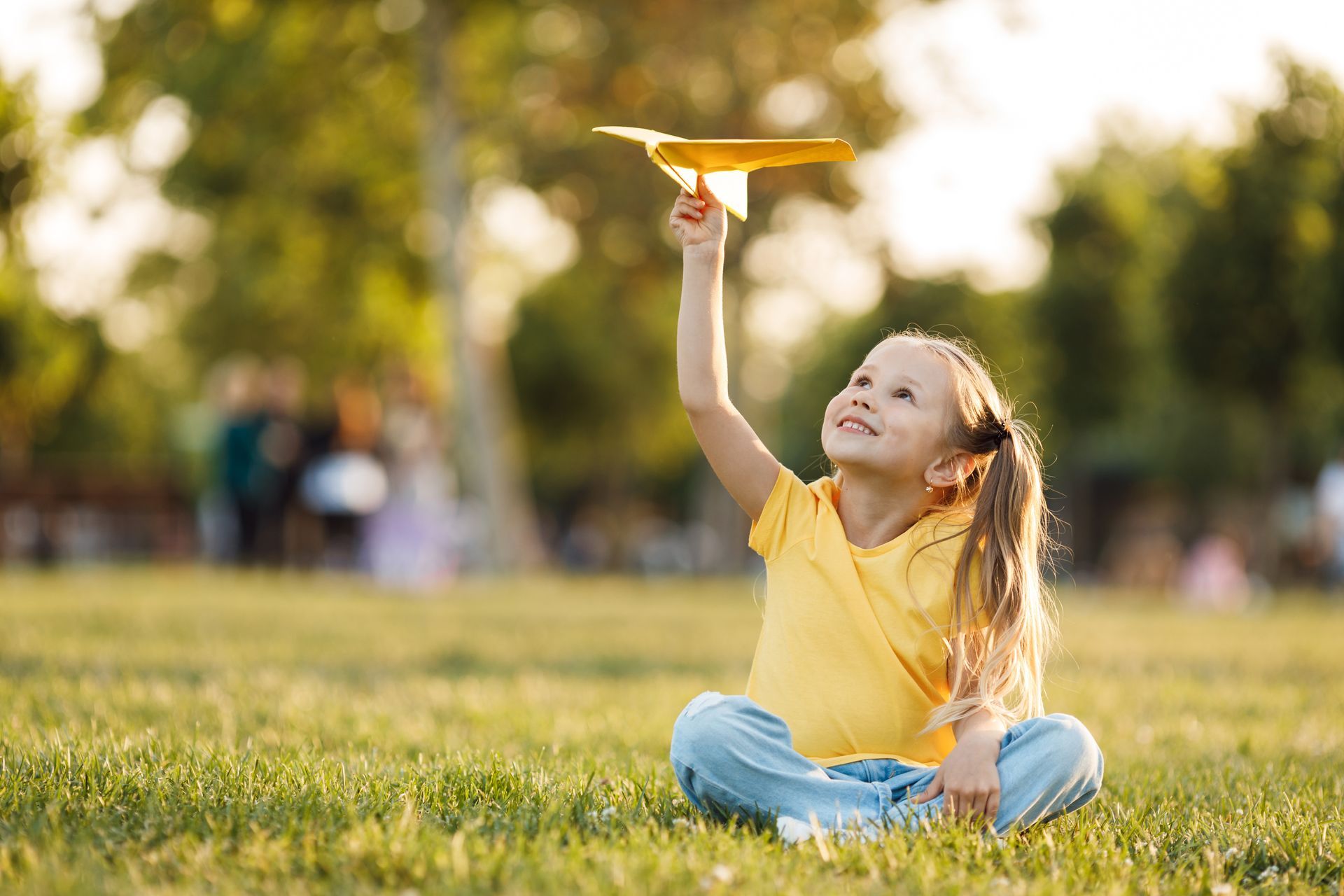 Child in a yellow shirt sitting on grass, smiling up at a toy airplane in a sunny park.