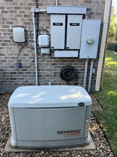 A Generac home generator installed outdoors on a stone base next to electrical boxes on a brick wall.