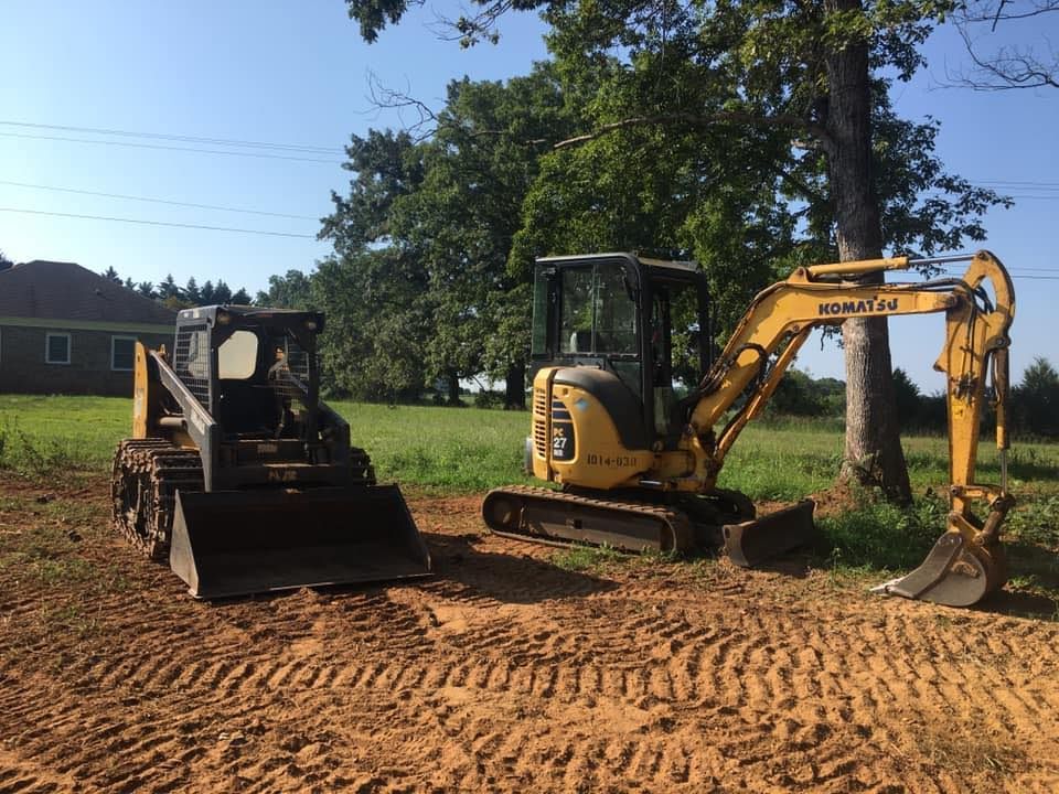 A skid steer and a yellow excavator parked on a dirt path in a grassy field.