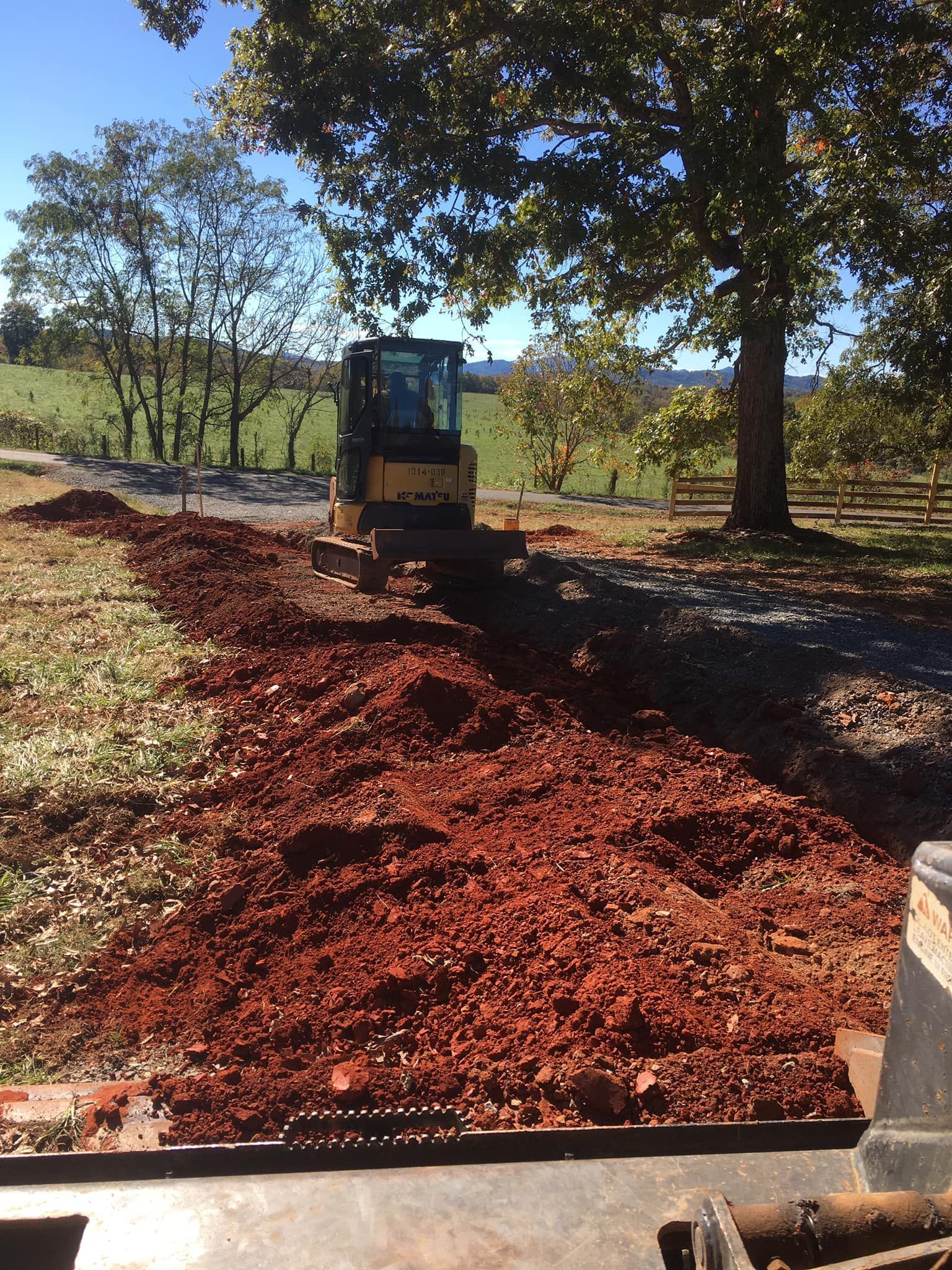 Mini excavator digging a trench along a dirt path next to a field. Sunny day.