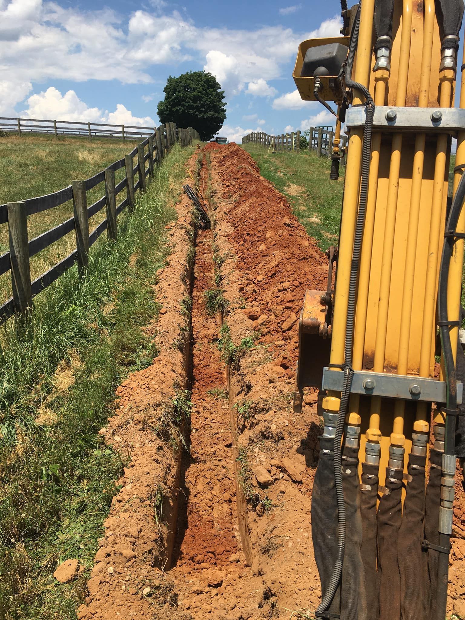 A trench dug in a grassy field, next to a wooden fence. Yellow excavator arm visible. Blue sky.
