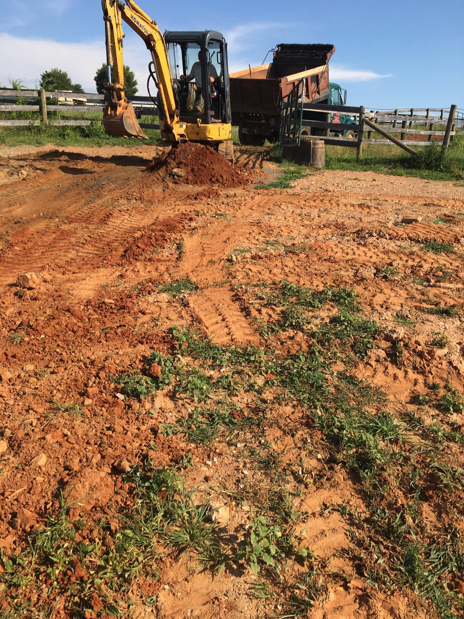 Mini excavator digging dirt, dump truck in background on a sunny day.