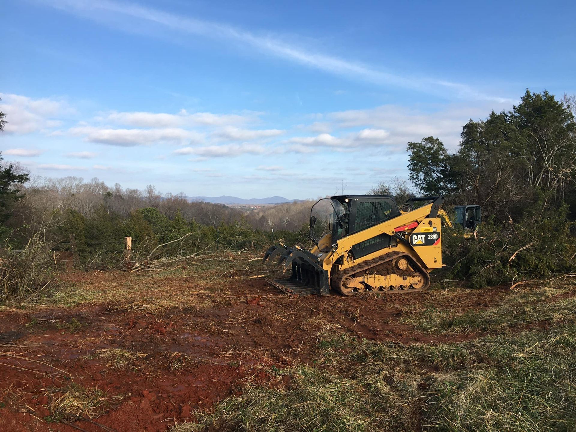 Yellow skid steer clearing land with brush cutter, brown earth, trees, blue sky.