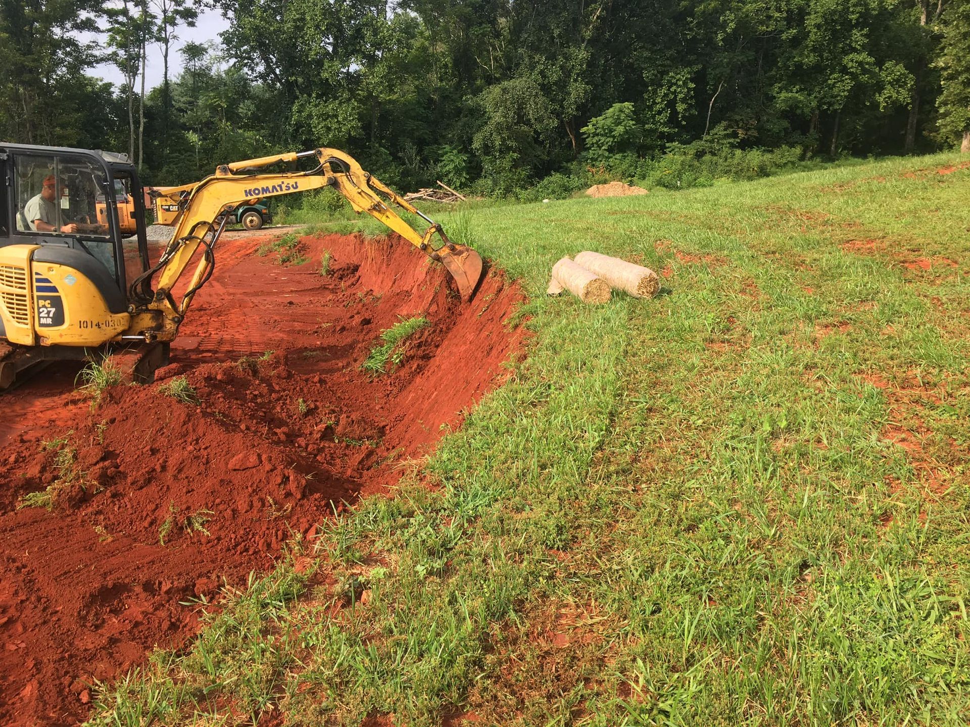 Yellow excavator digging into red soil on a grassy hillside, trees in background.