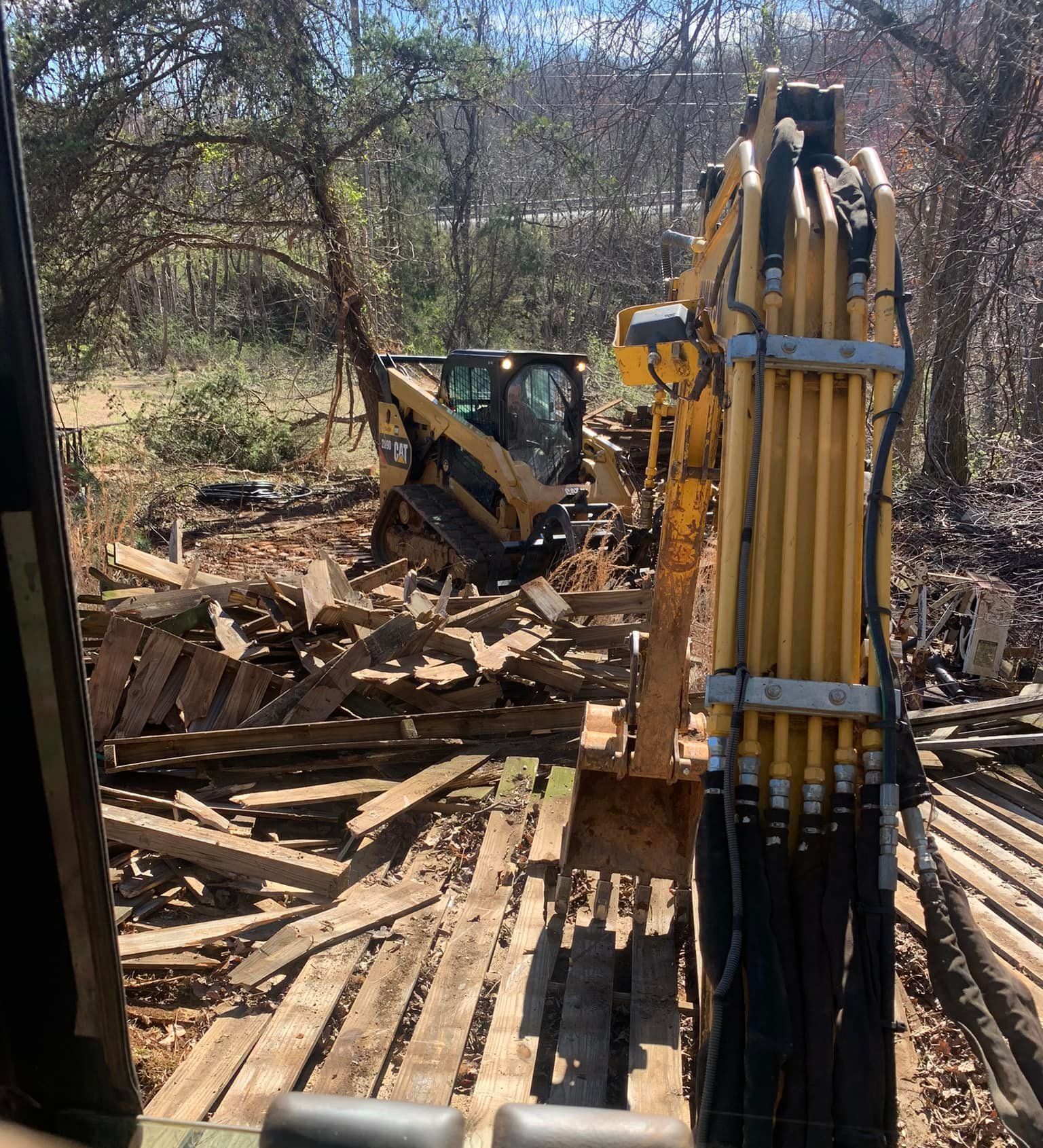 Excavator demolishing a wooden structure with a bulldozer in the background, in a wooded area.