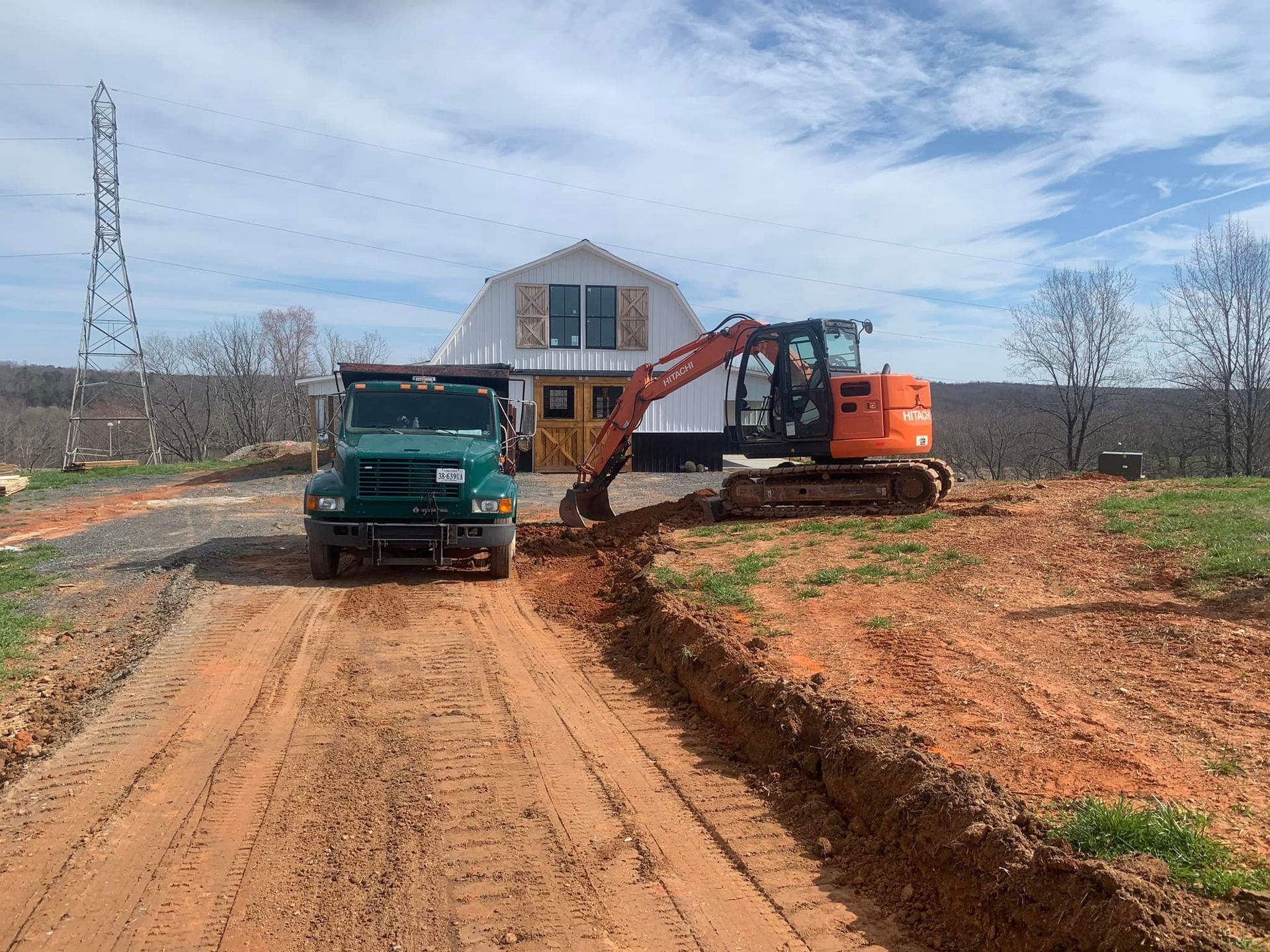 Construction scene: Excavator digging near a white barn, dump truck on dirt road.
