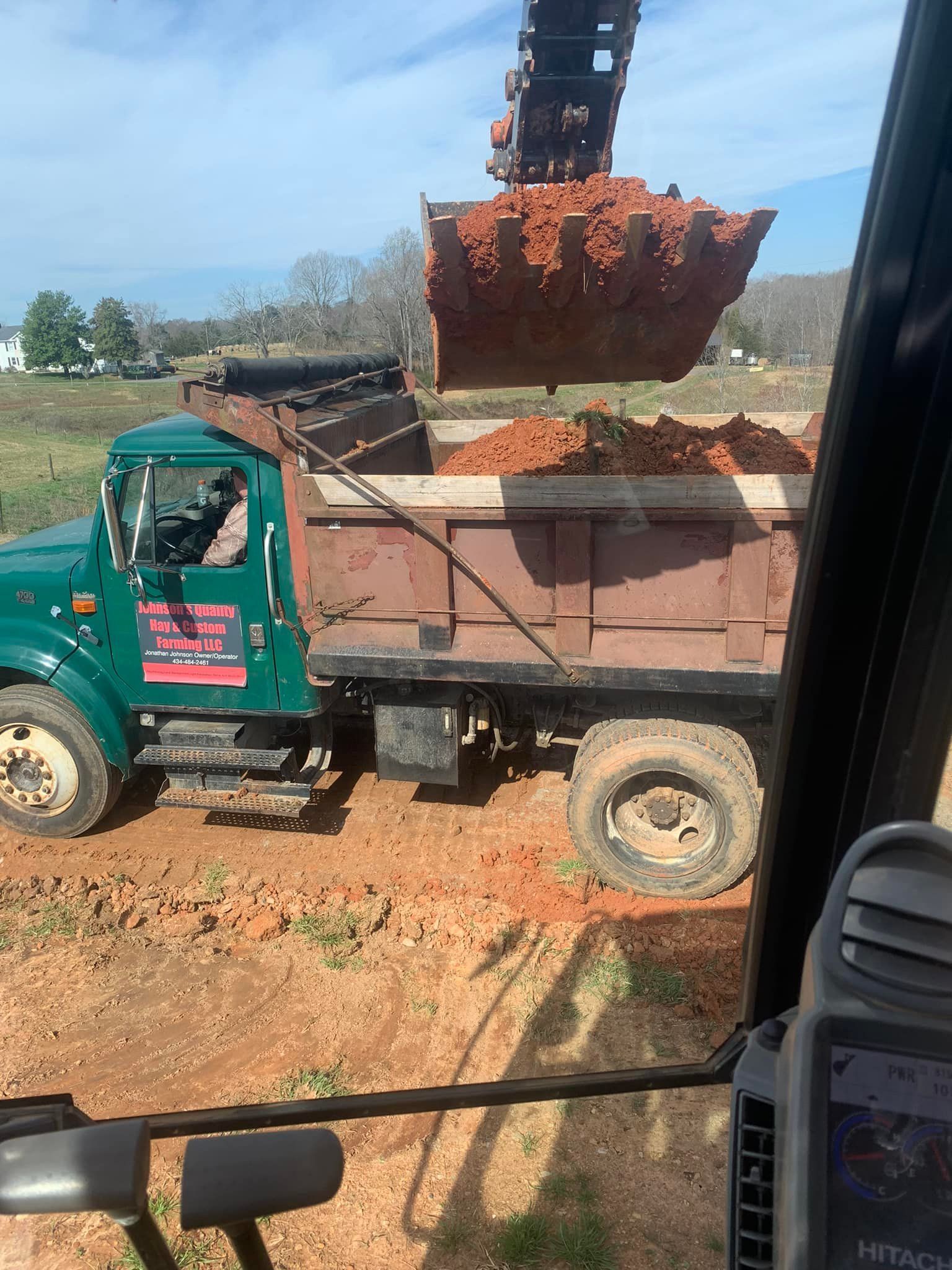 Excavator dumping red dirt into a green dump truck, on a sunny day.