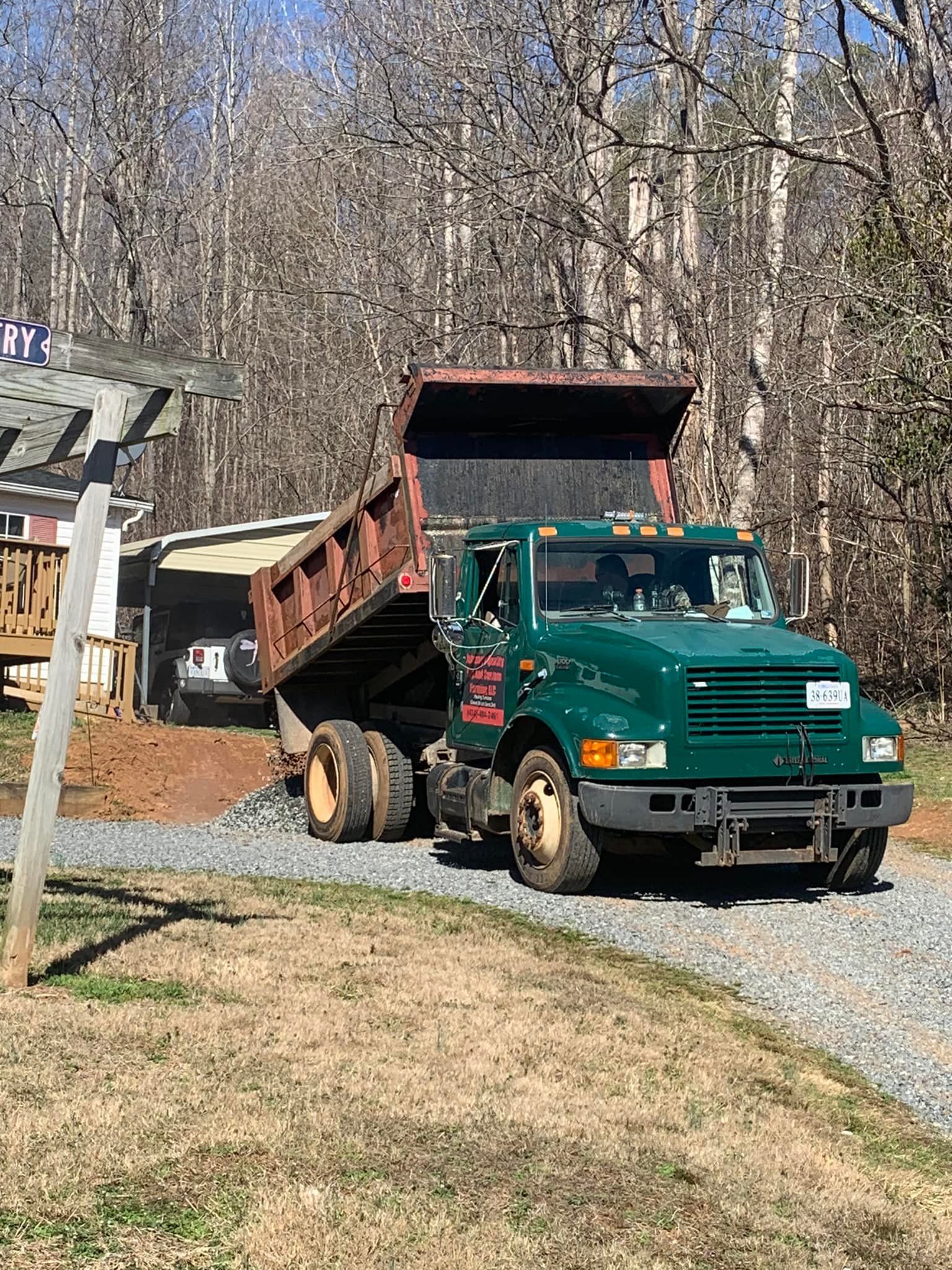 Green dump truck unloading material on gravel driveway, near a house and trees.