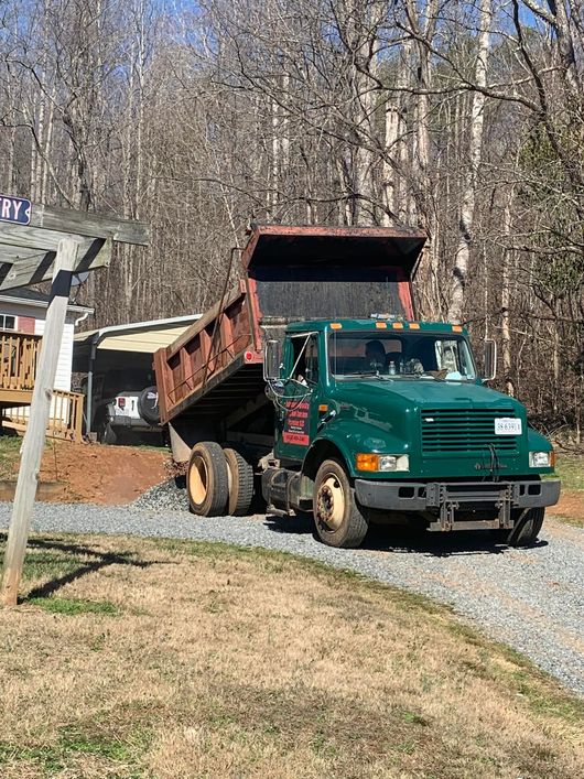 Green dump truck unloading dirt on a gravel driveway next to a house with a wooden porch.