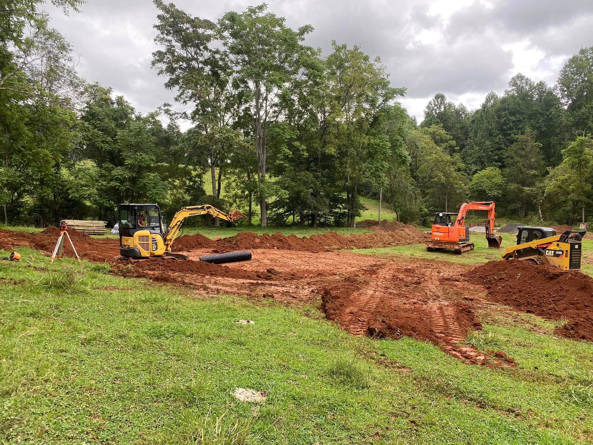 Excavators on a grassy field, digging in red soil, with trees in the background under a cloudy sky.