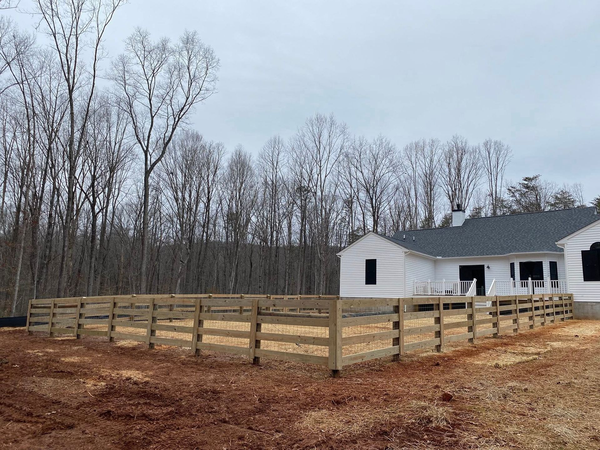 Wooden fence surrounding a white house under construction, with a backdrop of bare trees on a cloudy day.