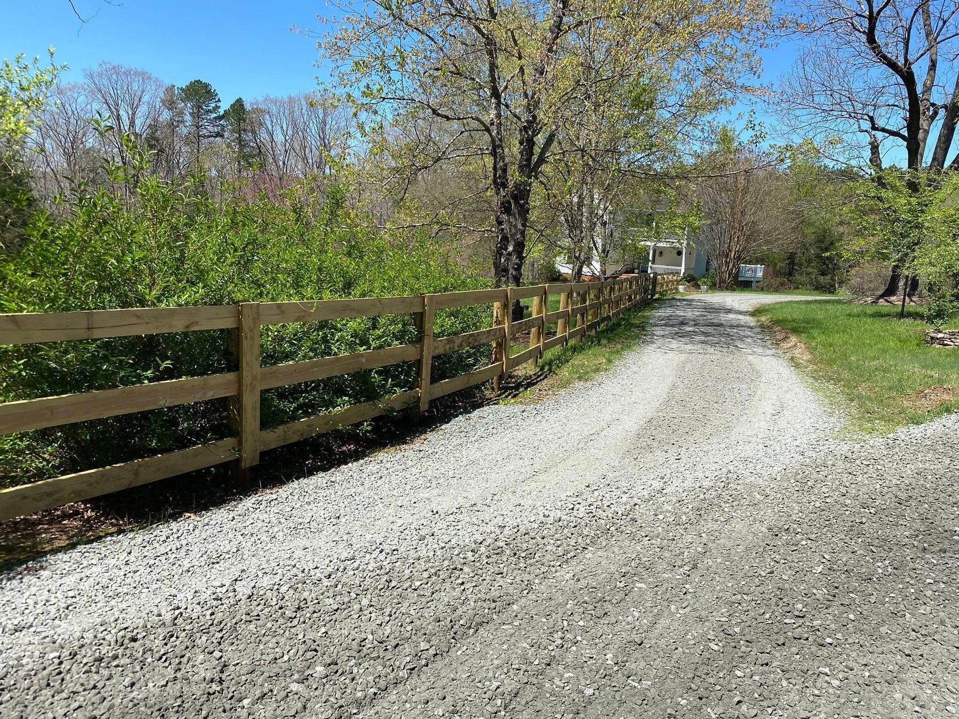 Gravel driveway with a wooden fence alongside a treeline on a sunny day.