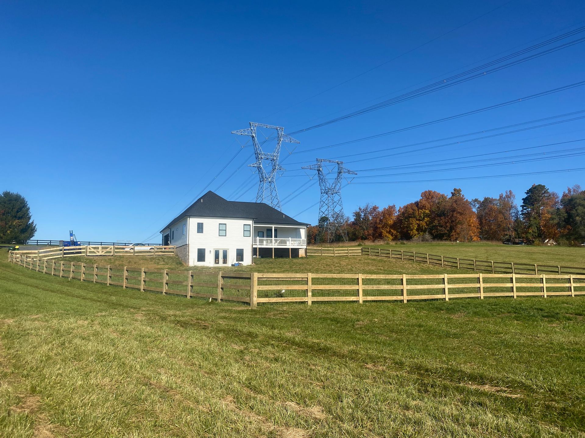White house with wrap-around porch and wooden fence in a field, power lines overhead, blue sky.