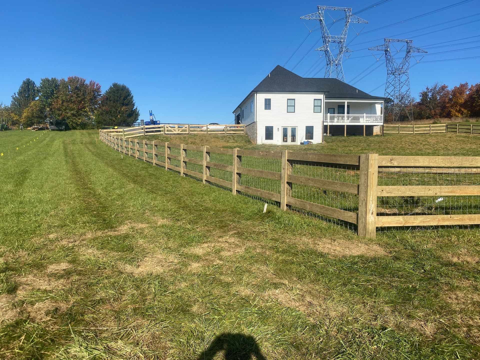 Two-story white house with wooden fence in a grassy field on a sunny day. Power lines visible overhead.