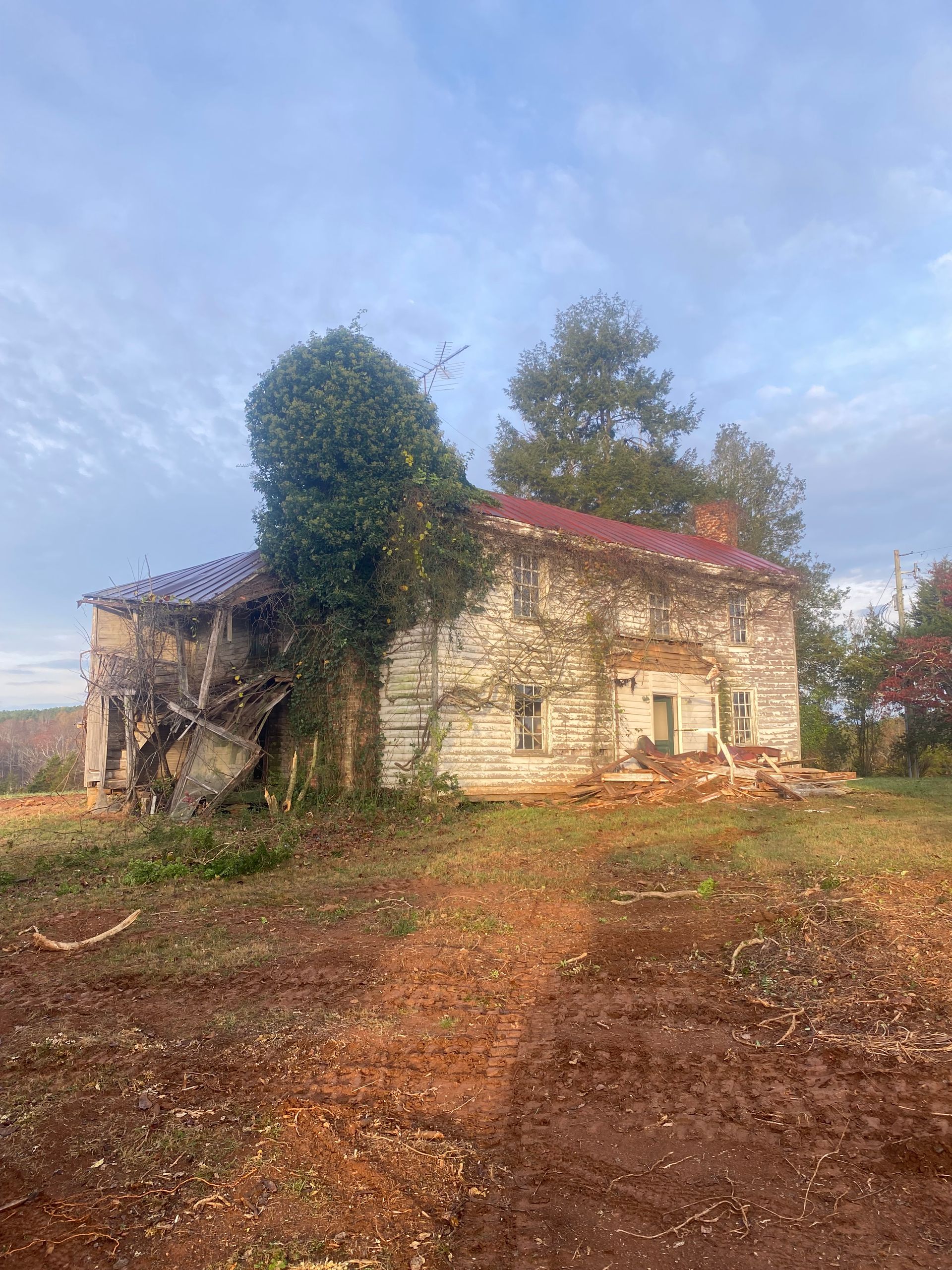 Dilapidated white building with vines, red roof, set on a brown earth. Cloudy sky.