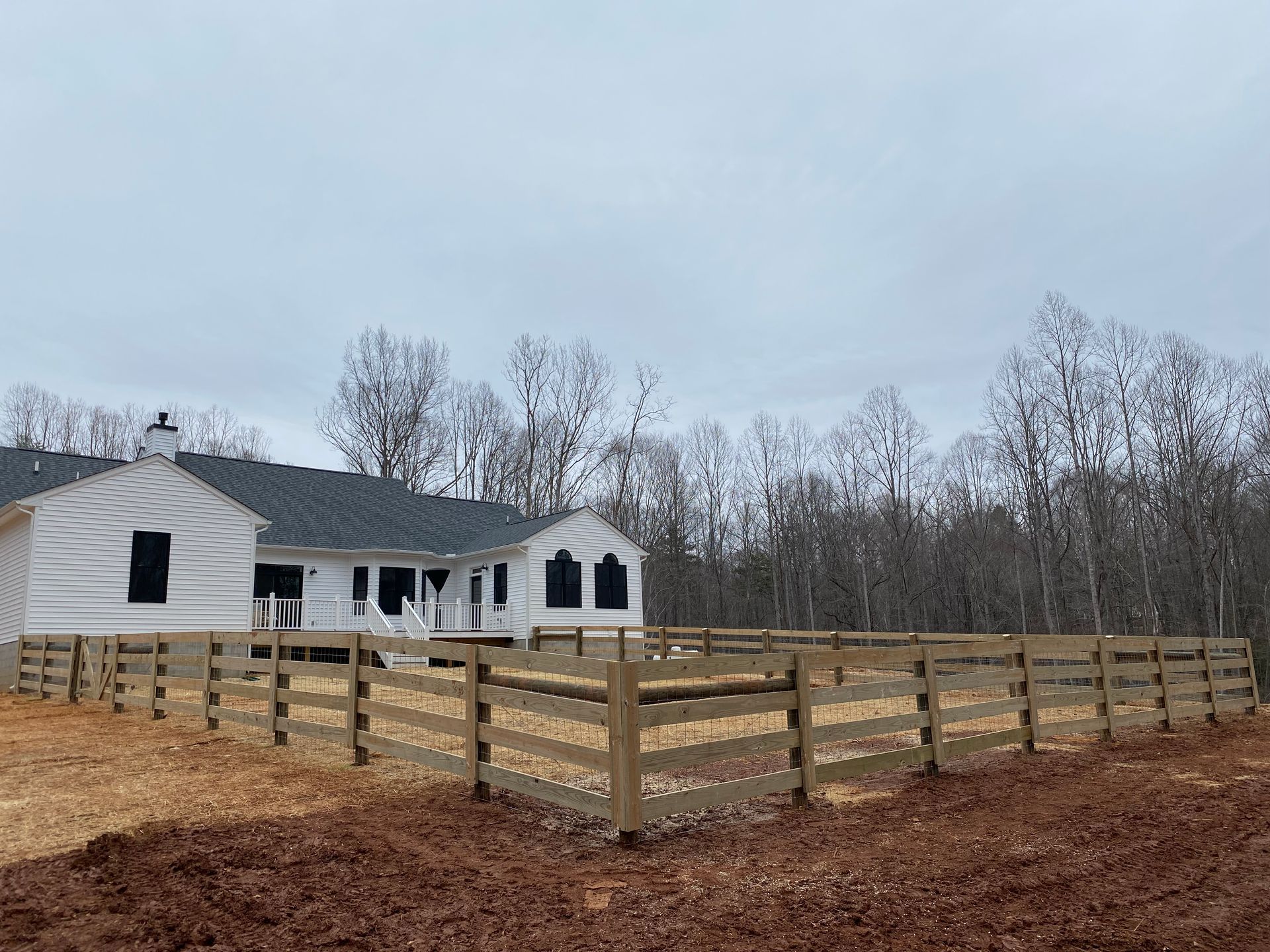 A new house under construction with a wooden fence in front, surrounded by brown leaves and trees on a cloudy day.