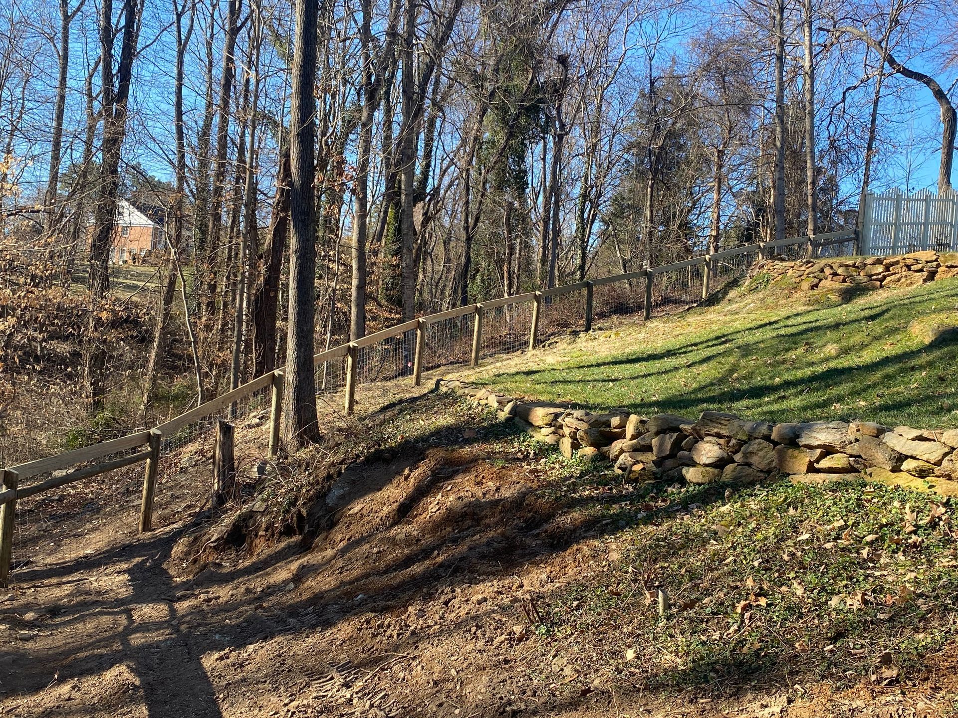 A dirt path winds through a hillside with a wooden fence. Trees line the background with a grassy area.
