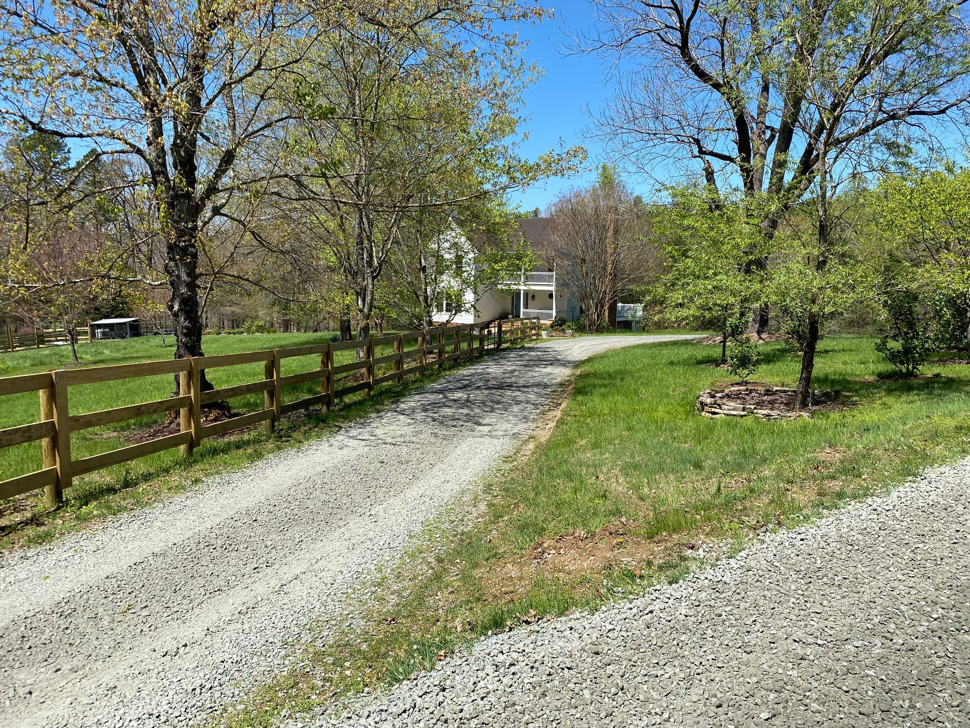 Gravel driveway leading to a white house, flanked by a wooden fence and grassy fields under a blue sky.