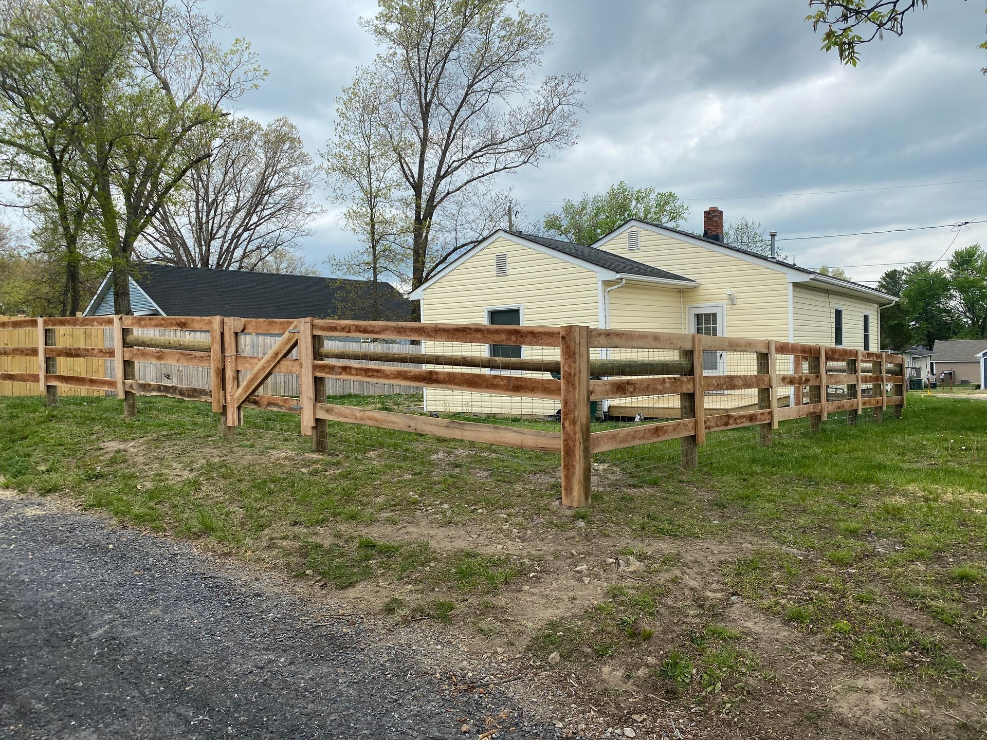 Wooden split-rail fence surrounds a yellow house on a grassy lot, under a cloudy sky.