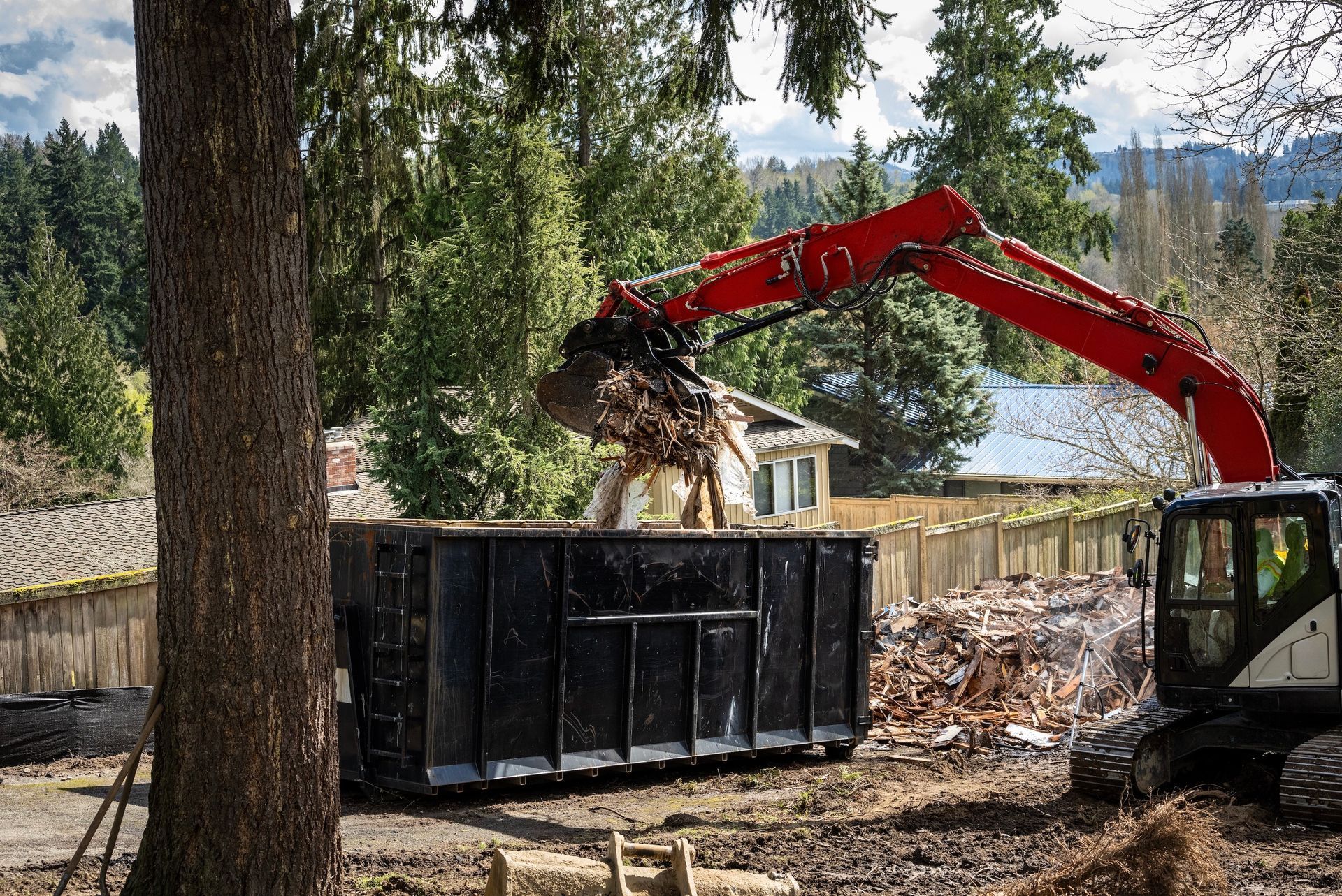 Red excavator loading debris into a black dumpster near a house.