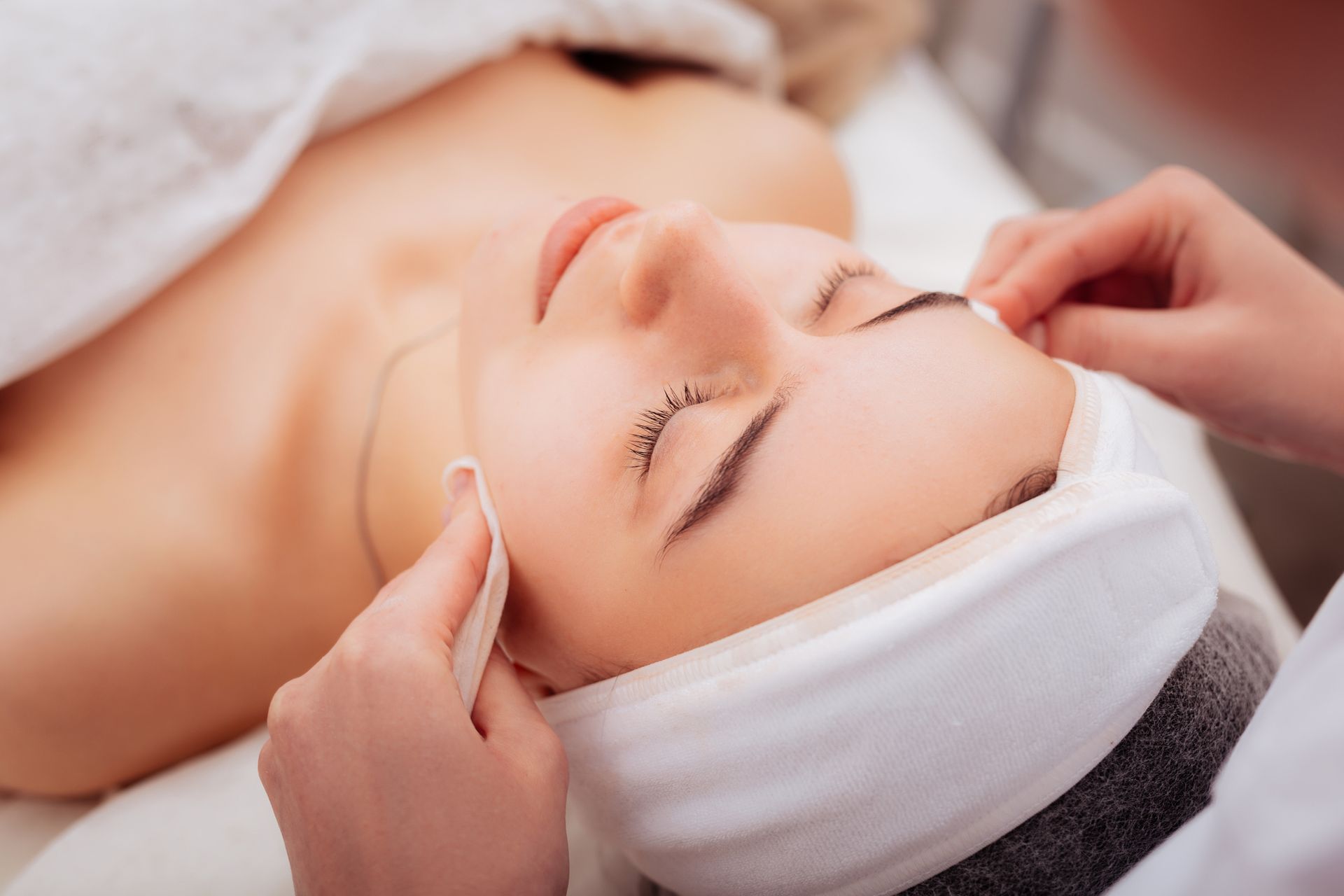 Woman receiving facial treatment, lying on a bed with closed eyes, hands holding cotton pads.