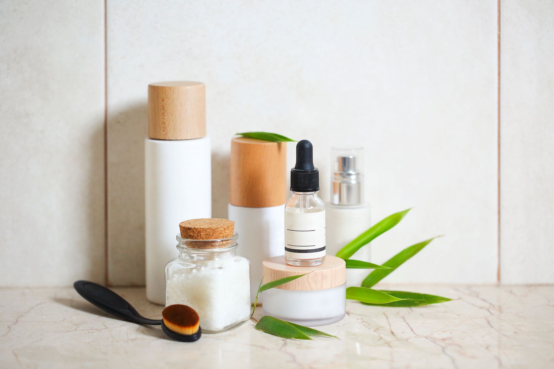 Skincare products: bottles, jars, dropper, and a makeup brush with green leaves on a beige surface.