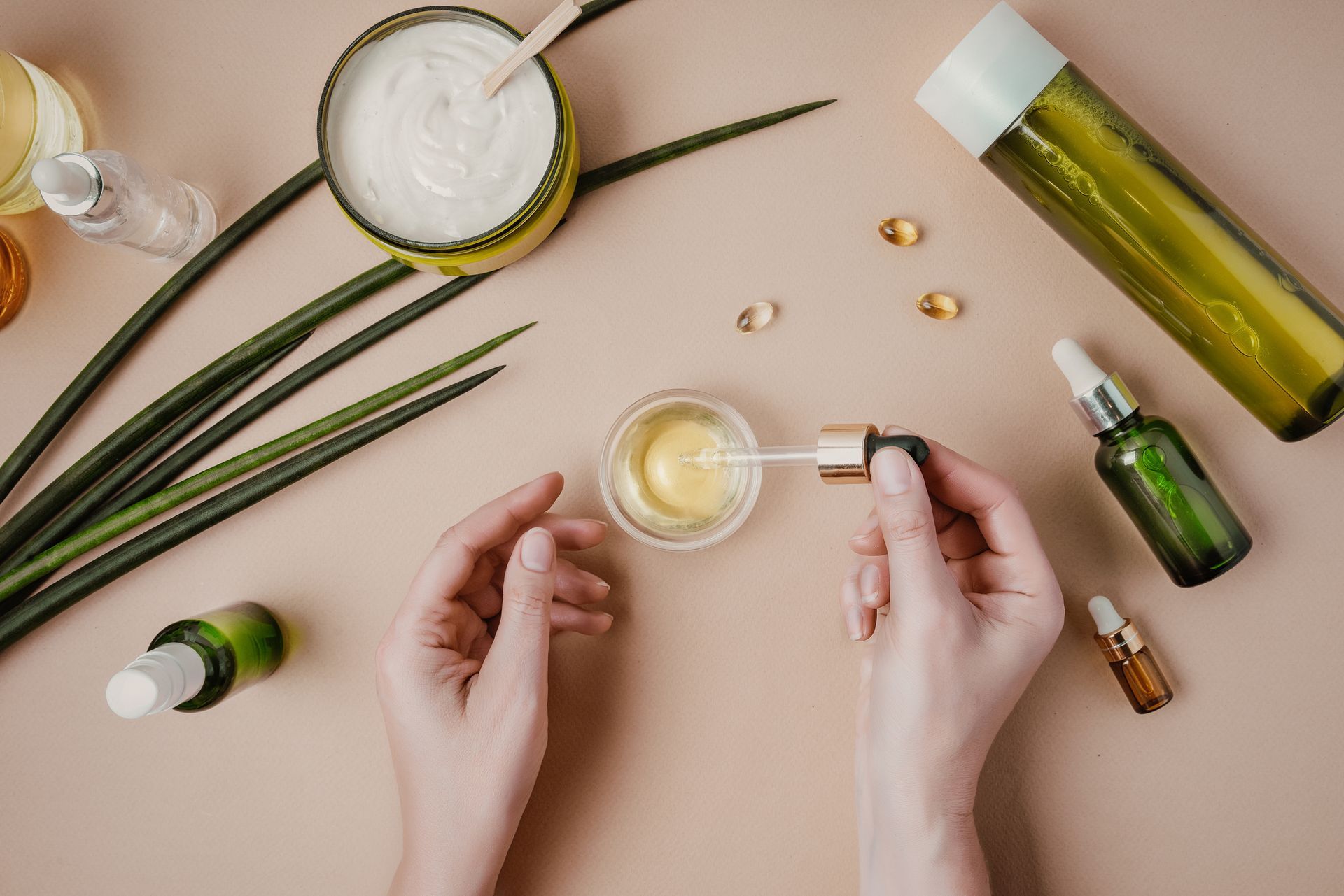 Hands holding dropper over small bowl with beauty products on beige surface.