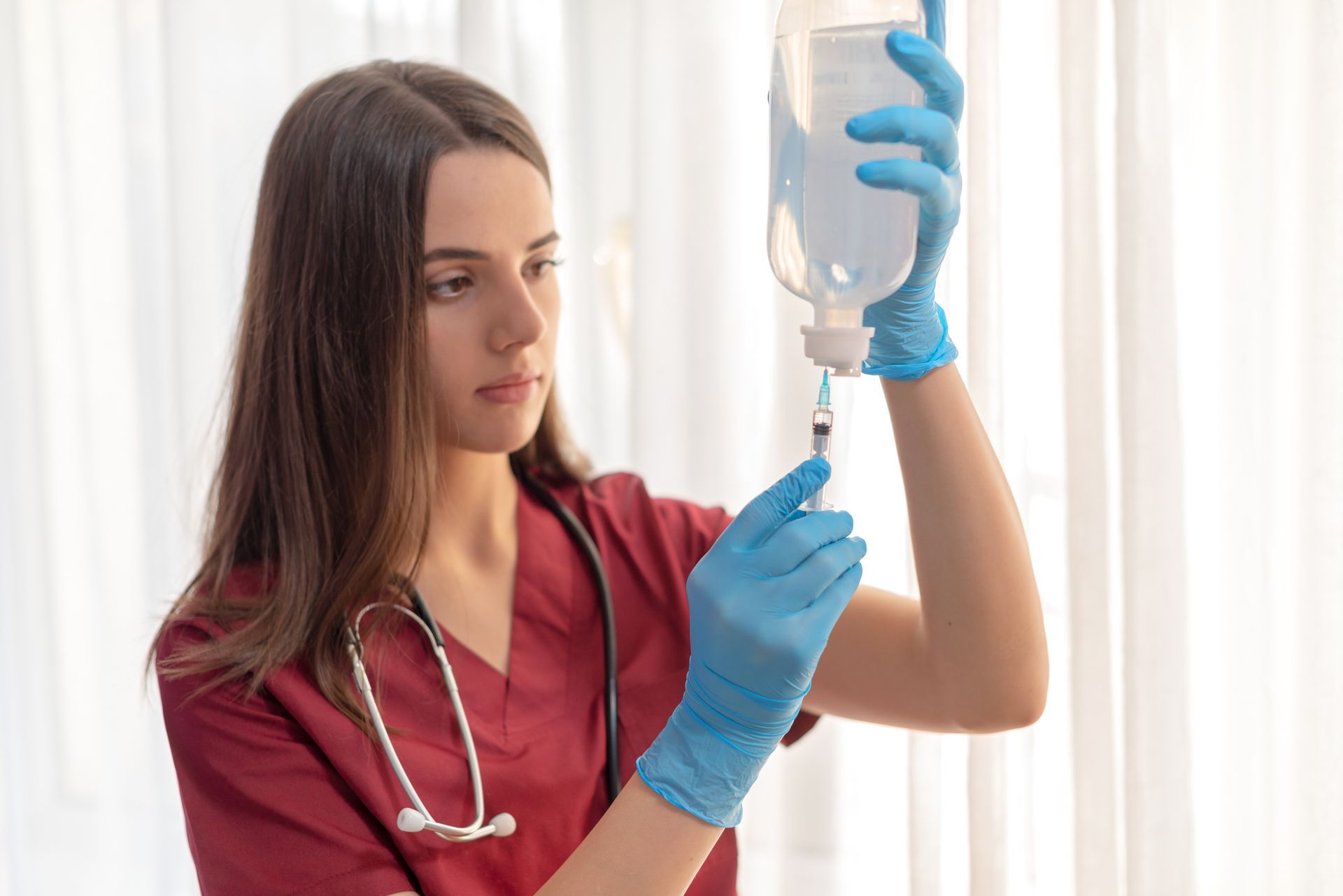 Nurse in maroon scrubs with stethoscope preparing IV drip in a bright room.