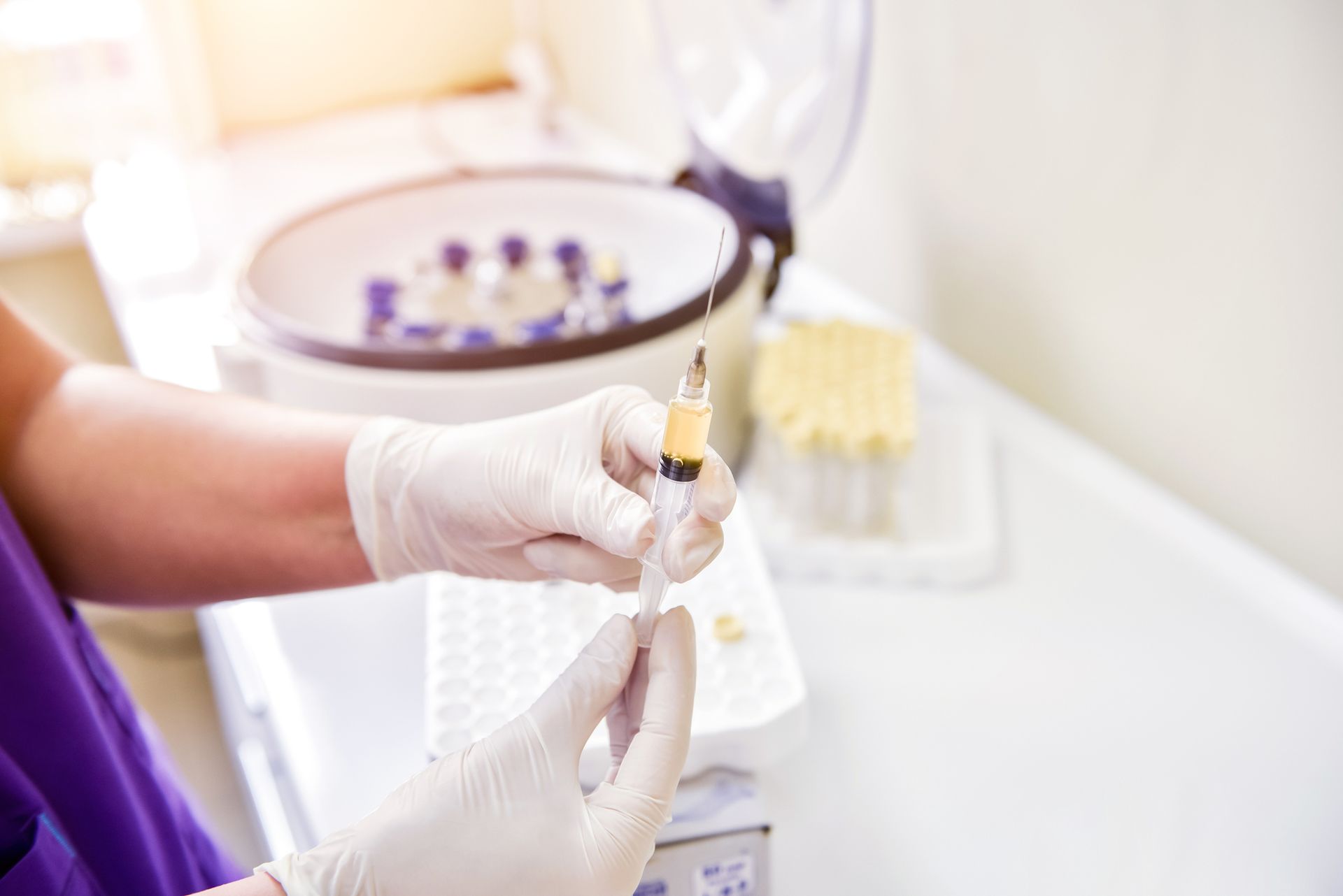 Person in gloves holding syringe, filling it with yellow liquid. Centrifuge machine and vials in background. Person in gloves holding syringe, filling it with yellow liquid. Centrifuge machine and vials in background.