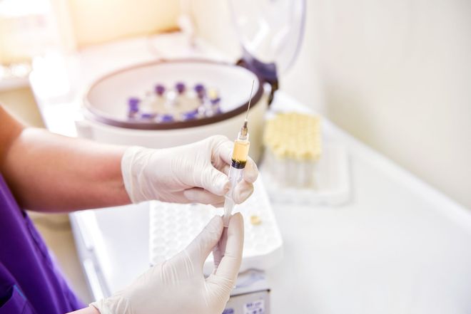 Person in gloves holding syringe, filling it with yellow liquid. Centrifuge machine and vials in background.